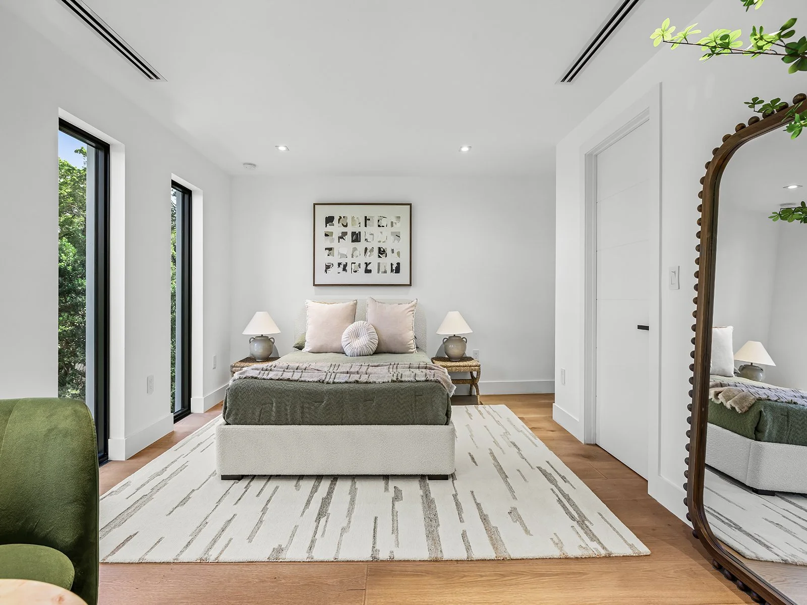 Modern bedroom with white walls, hardwood floor, bed with beige headboard, multiple pillows, two nightstands with lamps, large mirror, and three tall windows with green trees outside.