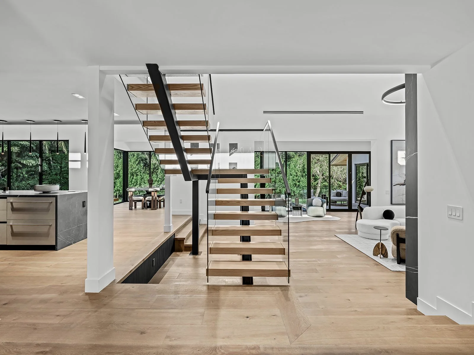 Modern open-concept home interior with wooden stairs, white walls, large windows, and minimalistic furniture.