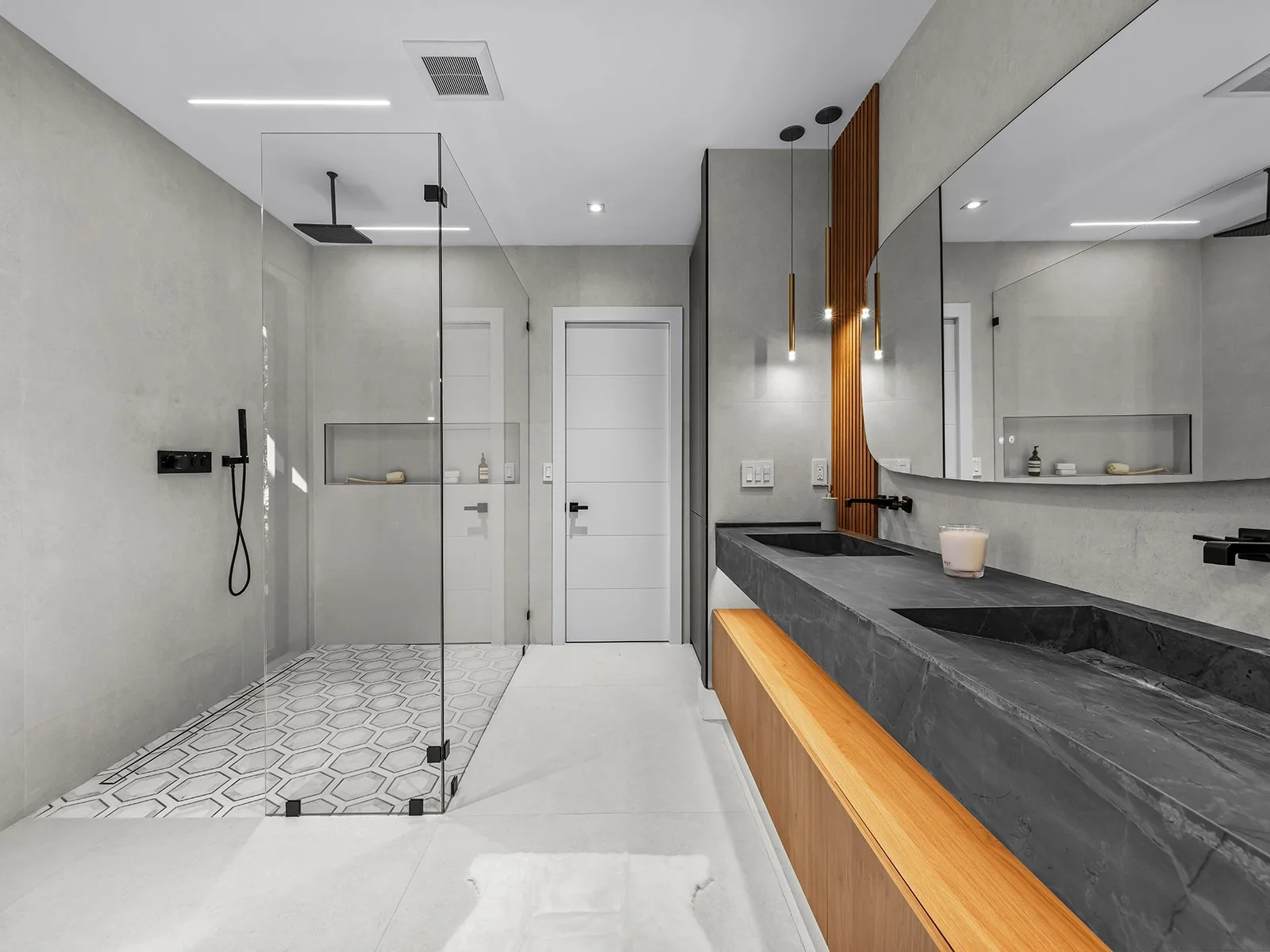 Modern bathroom with a walk-in glass shower, double black sinks on a dark stone vanity, and large mirror. Features include pendant lights and wood accents.