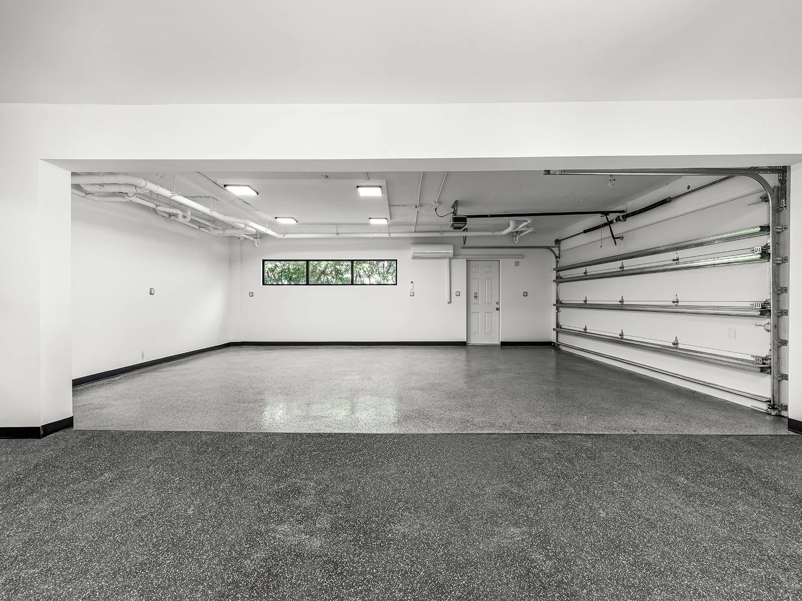 Empty garage with white walls, black baseboards, a gray speckled floor, a closed garage door, a window, a door, and ceiling lights.