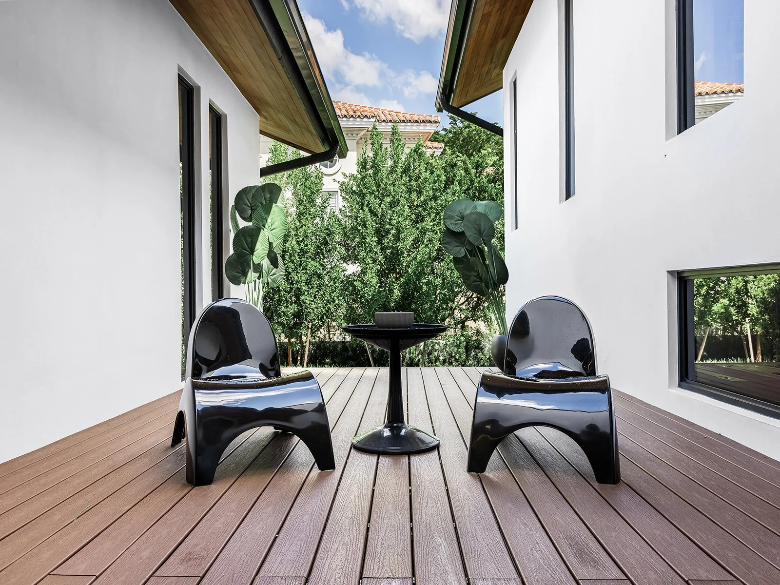 Modern outdoor patio with wooden decking, two black glossy chairs, a small black table, and lush green trees in the background, surrounded by white walls and windows.