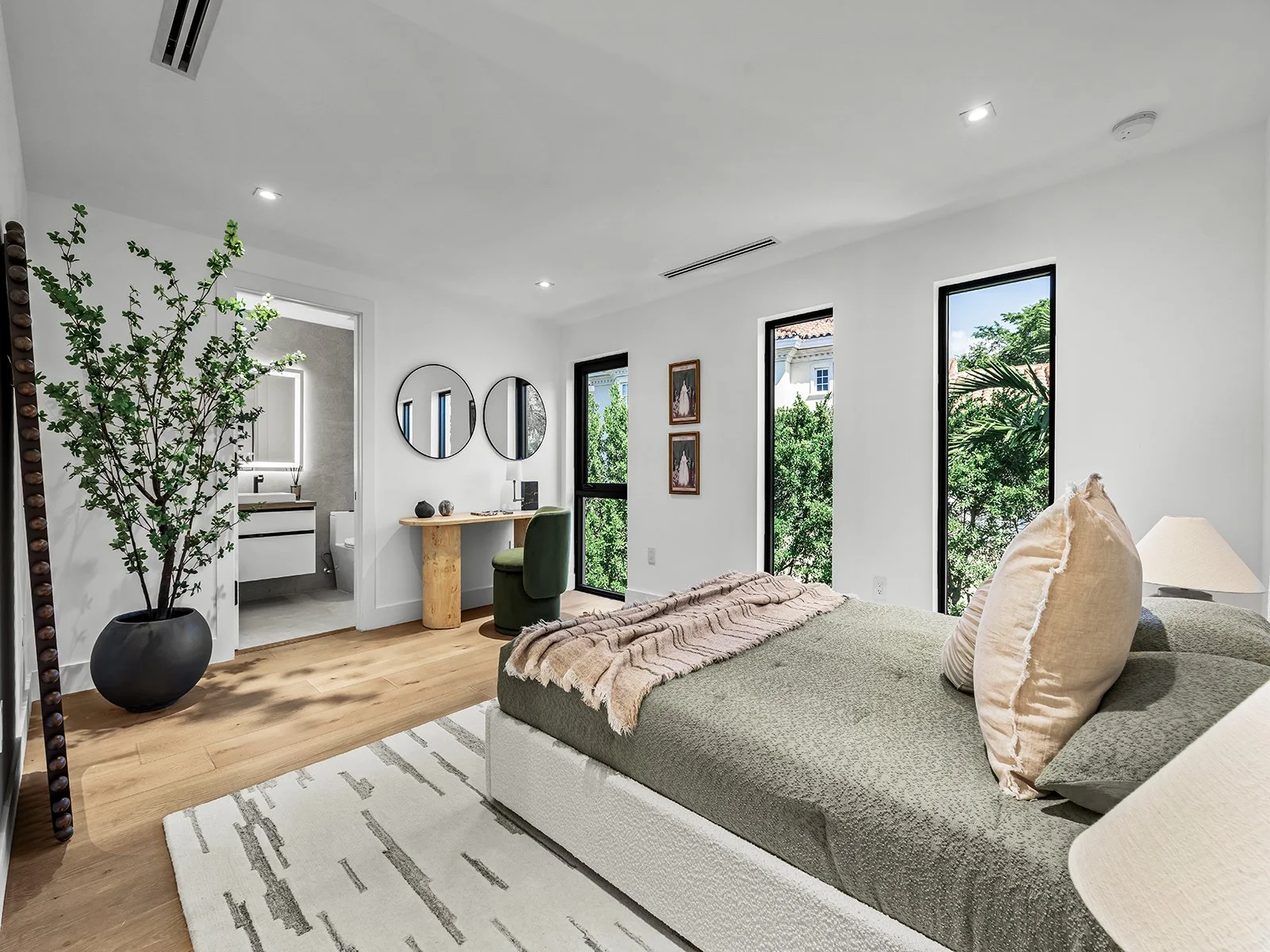Modern bedroom with large windows, neutral-colored bed, and decorative pillows. Features a white area rug on wooden flooring, a large potted plant, a dressing table with mirrors, and a view of greenery outside.