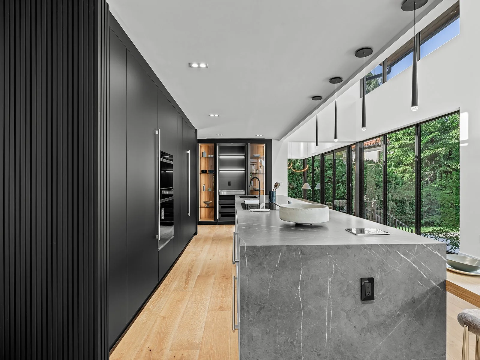 Modern kitchen with black cabinets, marble island, large windows with greenery view, wooden flooring, black pendant lights.