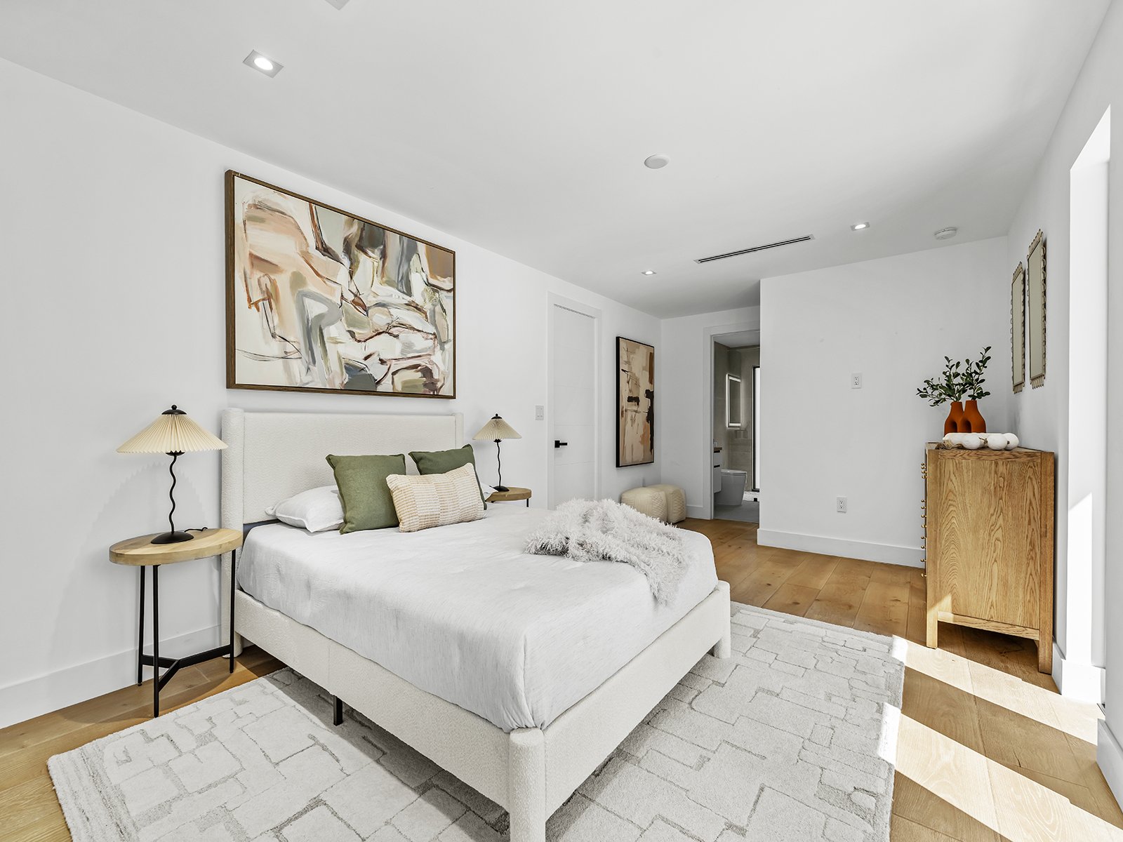 Modern white bedroom with a bed, abstract art above, two side tables with lamps, a wooden dresser with vases and greenery, and a view of a bathroom.