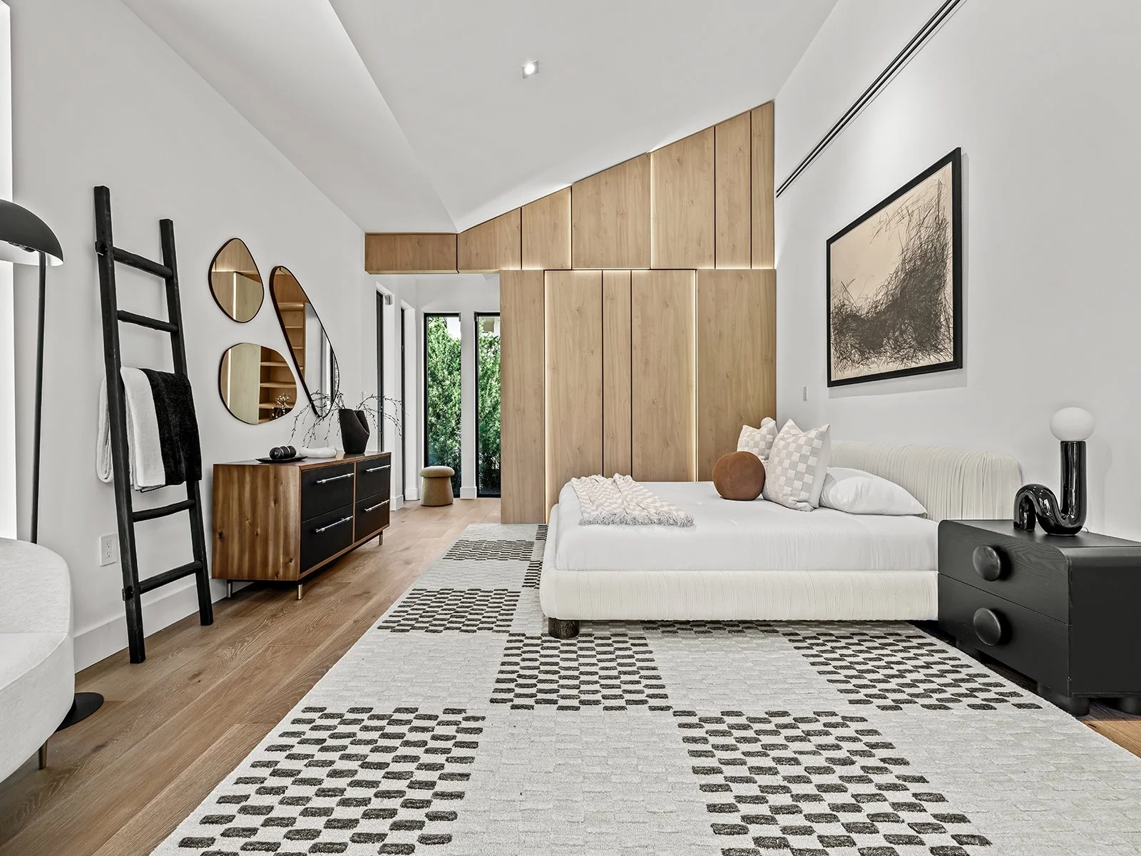 Modern bedroom with white walls, wooden accents, and black furniture. Features a bed with white linens and decorative pillows, a black nightstand with a lamp, and a black ladder with towels. Decorative mirrors and artwork adorn the walls. Light hardw