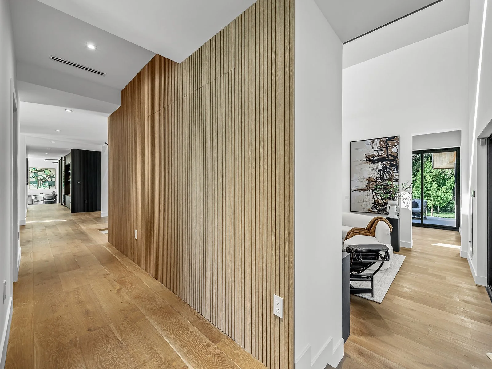 Interior of a modern home featuring a hallway with a wooden slat wall on the left and a living space with art, a chair, and large windows letting in natural light on the right
