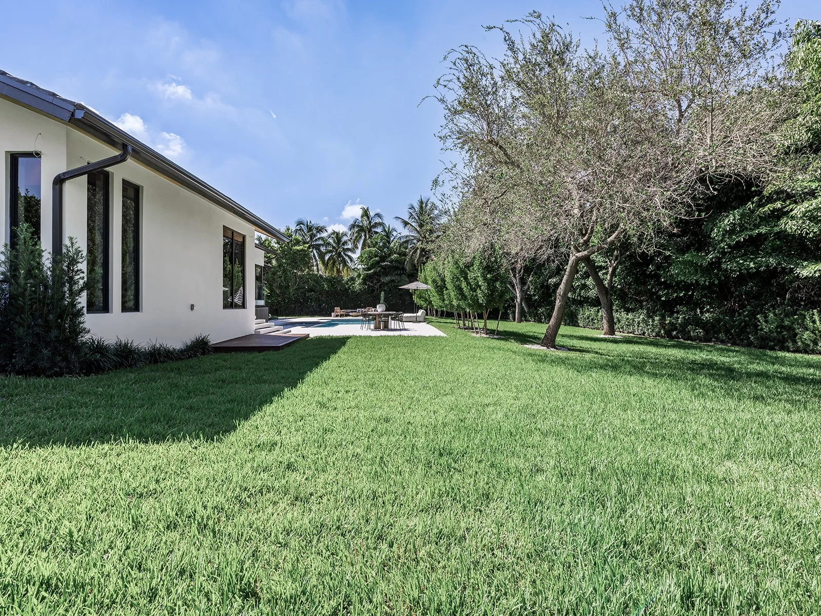 A modern white house with a backyard, green lawn, trees, and a patio with outdoor furniture, swimming pool, and umbrella under a sunny blue sky.