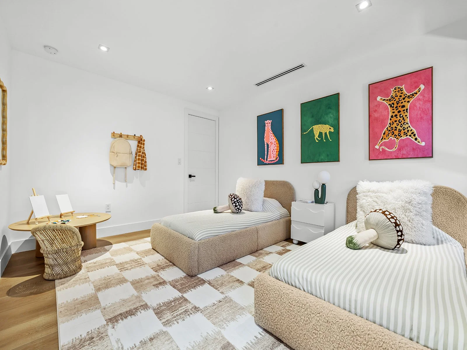 A modern bedroom with two single beds, each with beige and striped bedding and plush animal pillows, a white nightstand with a lamp, and three colorful animal-themed paintings on the wall. There is a small round table with a wicker chair and a wall h