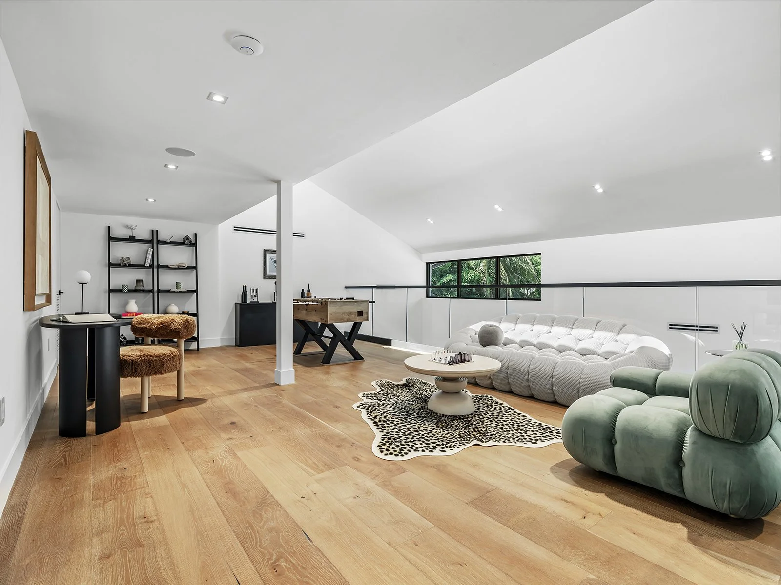 Modern living room with wooden floor, white walls, various contemporary furniture including a large white sofa, green armchair, small round table with chess, black shelf with decor, foosball table, and a window showing greenery outside.