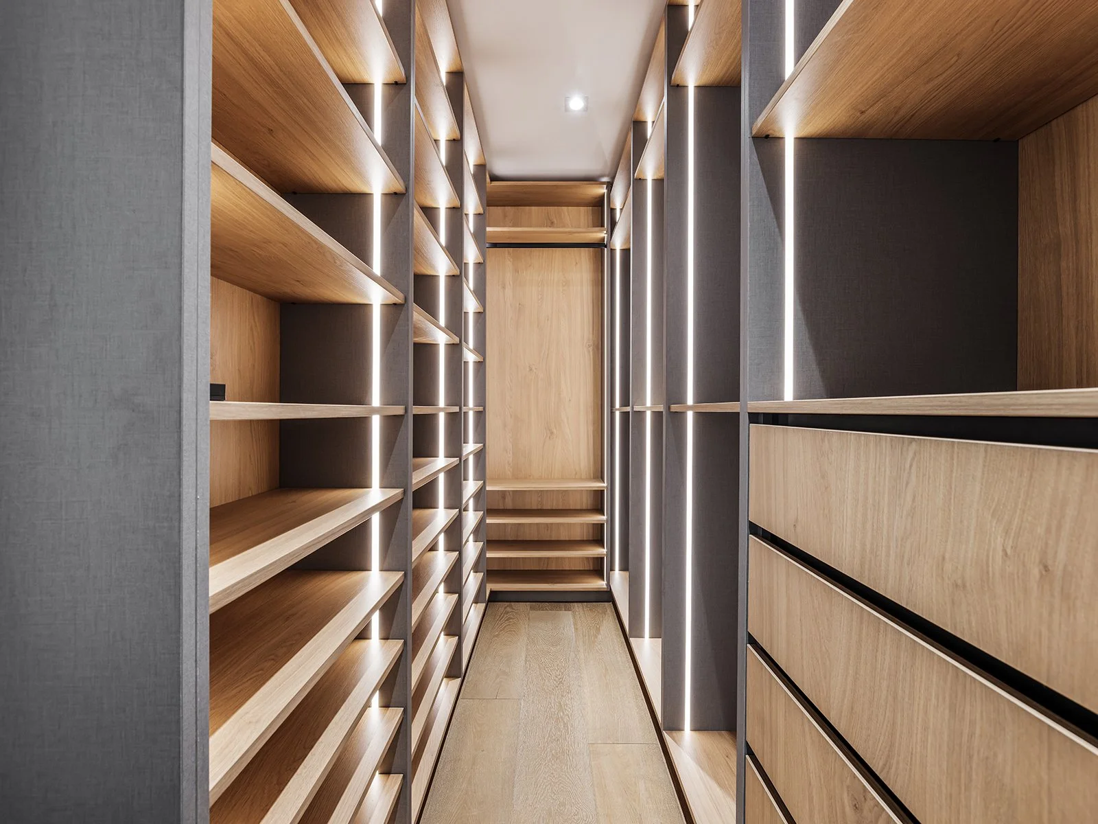 Empty walk-in closet with built-in wooden shelves and drawers, illuminated by vertical LED strip lights.