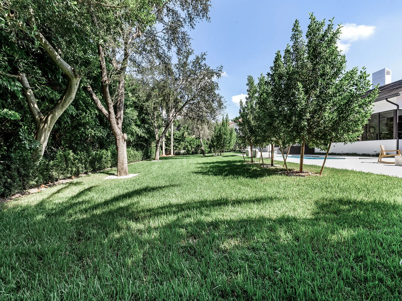 A lush green backyard with a well-maintained lawn, several trees on the left, and a modern house with a patio and swimming pool on the right, under a partly cloudy blue sky.