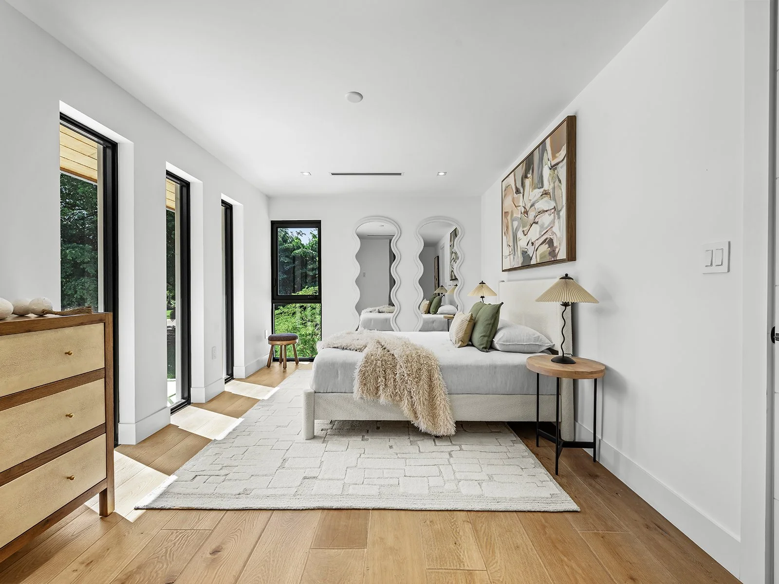 A modern bedroom with white walls, wooden flooring, and a large white bed with green and beige pillows. There are three tall narrow windows on the left, and a decorative mirror made of three wavy panels at the foot of the bed. A abstract painting han