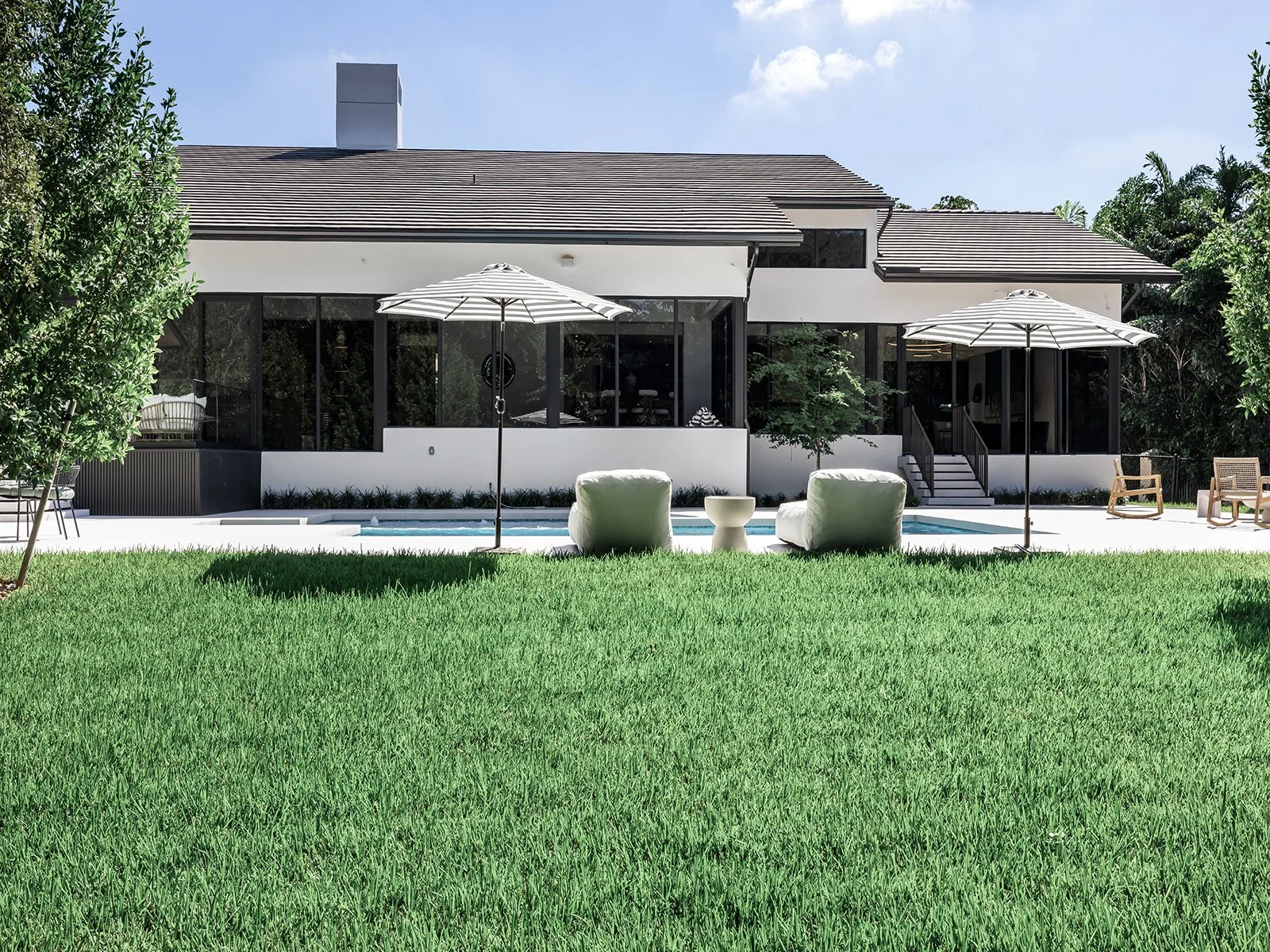 Modern house with large windows, outdoor seating with umbrellas, and a swimming pool surrounded by a lush green lawn.