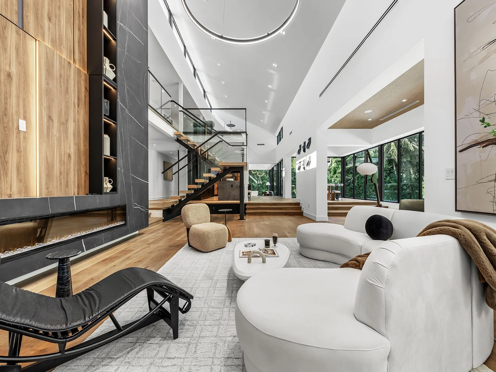 Modern living room with white curved sectional sofa, black lounge chair, beige pouf, and decorative objects on a coffee table, large windows with greenery outside, wooden flooring, contemporary artwork on the wall, and a staircase leading to an upper