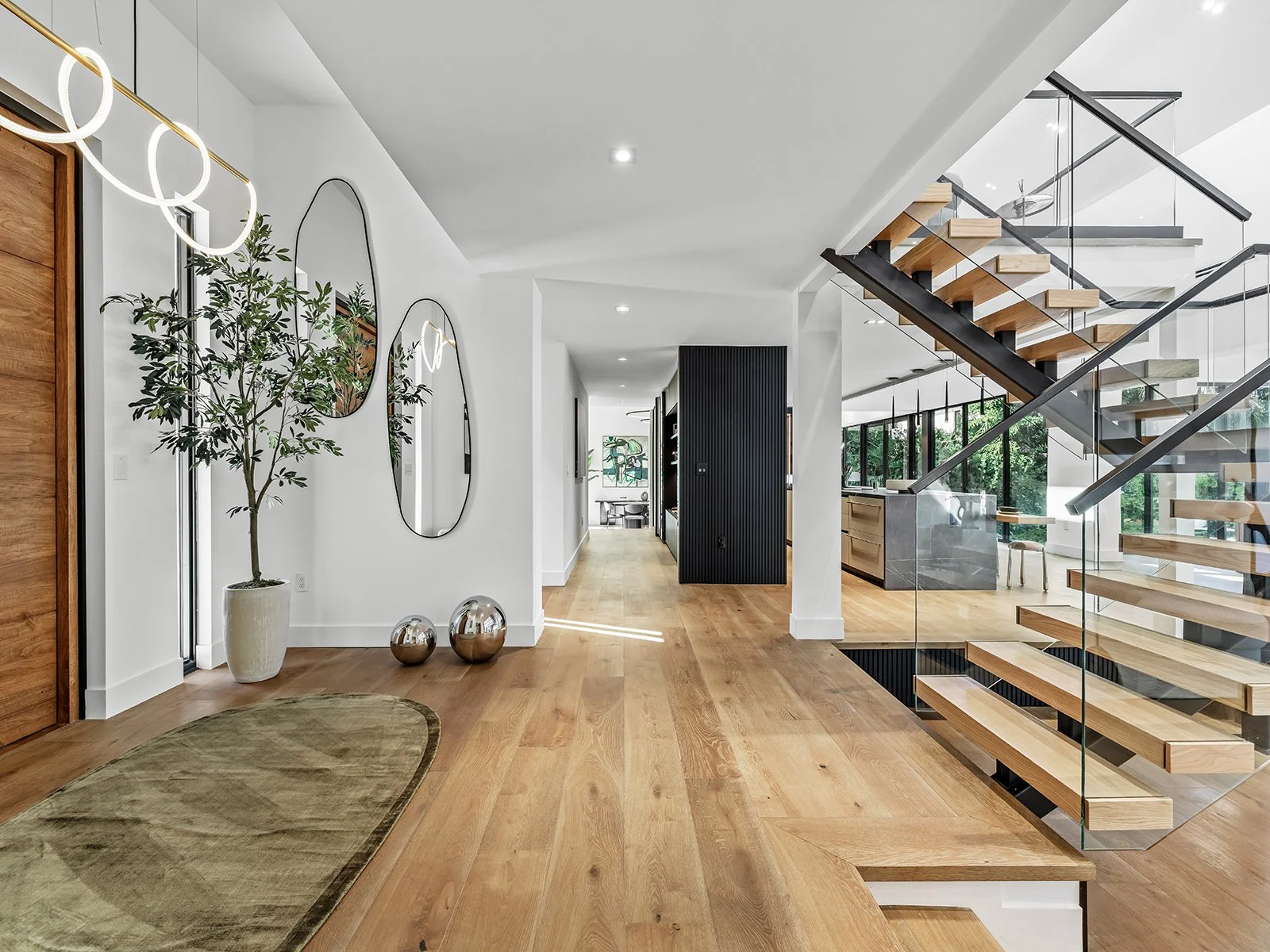 Modern, open-plan living space with wooden floors, a staircase with glass railing and wooden steps, large windows, and minimal furniture.