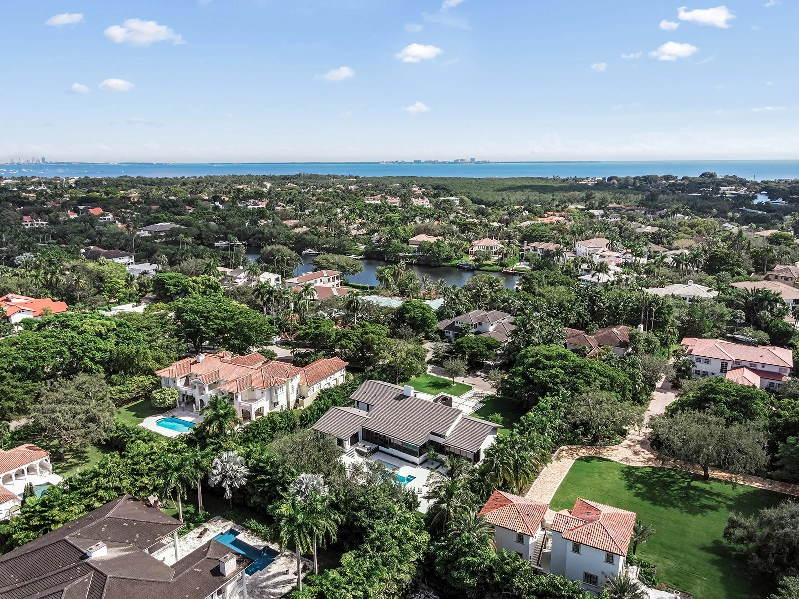 Aerial view of a suburban neighborhood with houses, lush trees, swimming pools, and a waterfront in the distance.