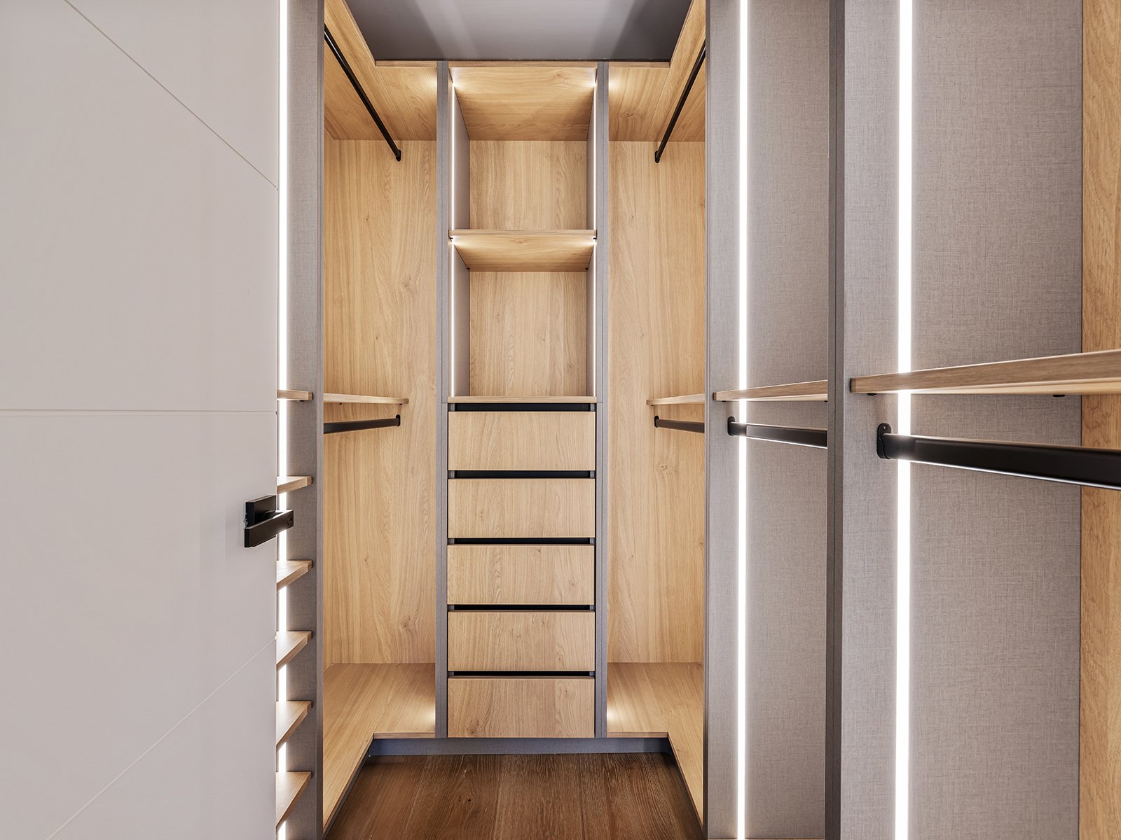 Empty walk-in closet with wooden shelves and tiled walls, illuminated by vertical LED lights.