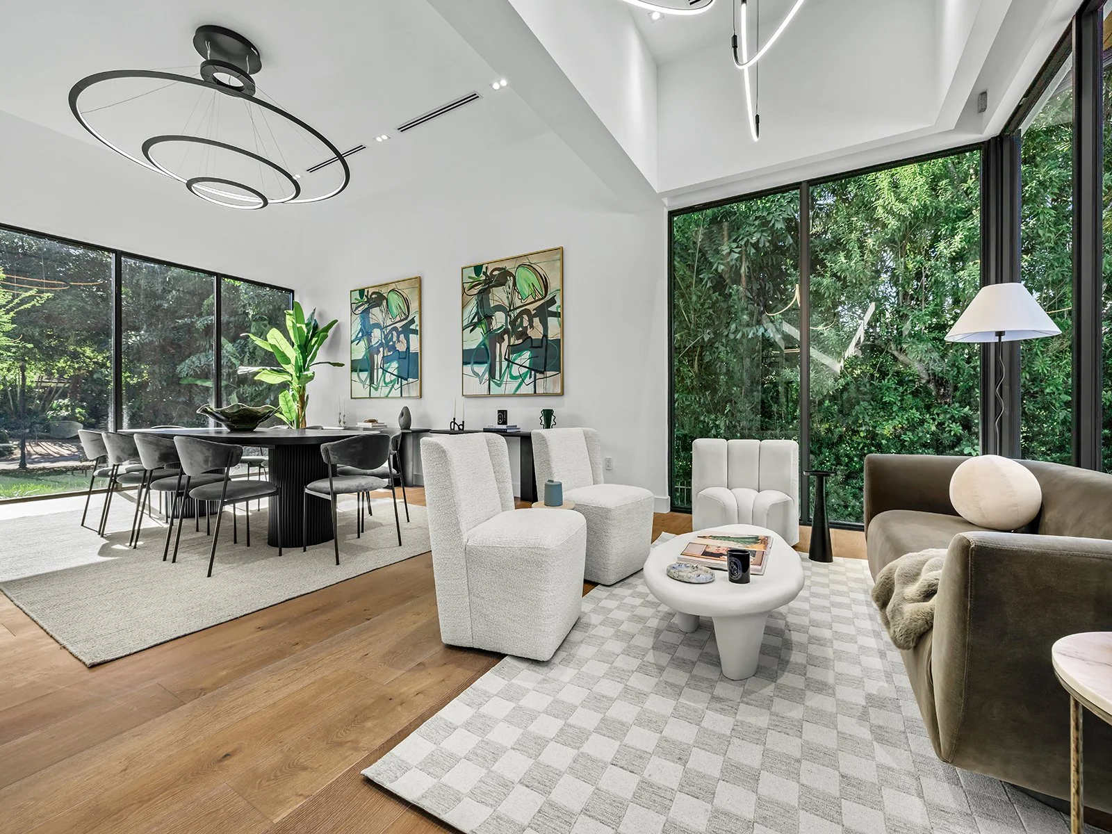 Modern living room with large windows, white walls, wooden flooring, white and beige furniture, abstract artwork, floor lamp, and greenery outside.
