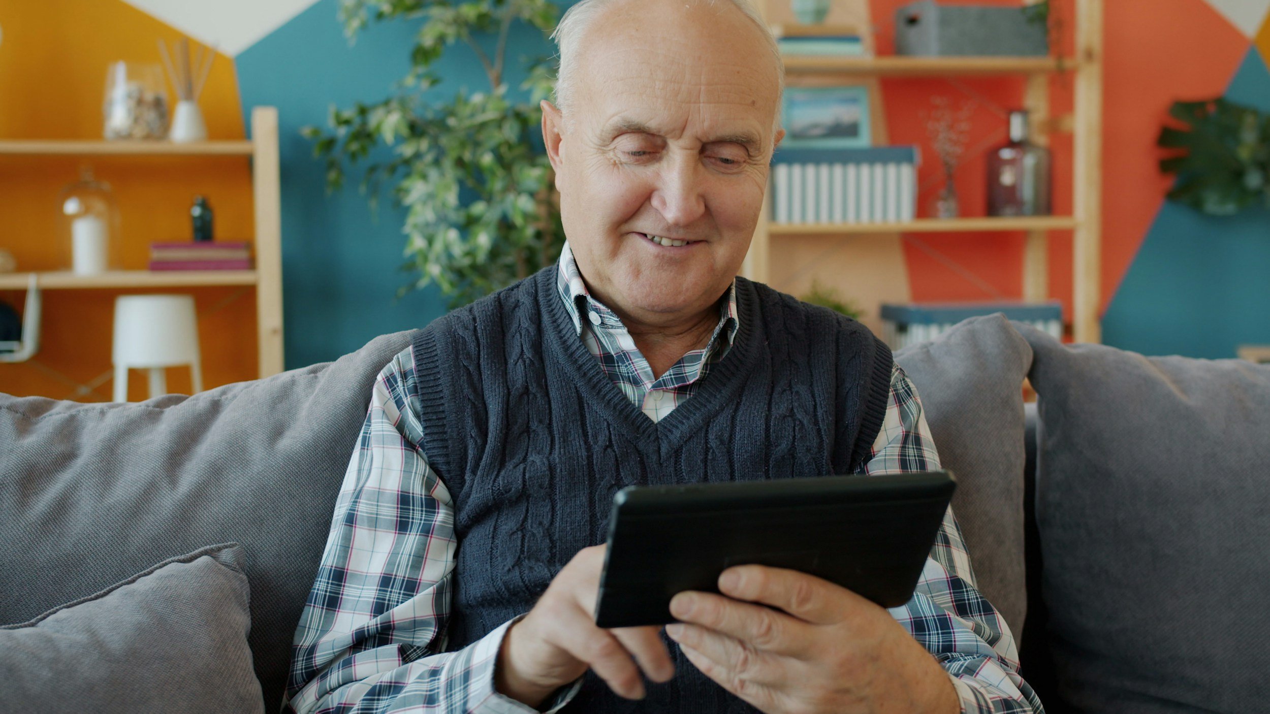 An elderly man sitting on a gray sofa, smiling as he looks at a black tablet in his hands in a colorful living room with bookshelves and a large potted plant in the background.