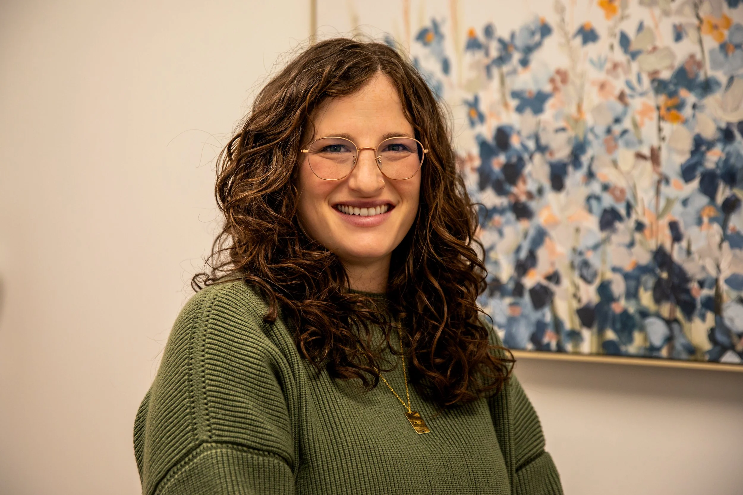 A woman with long curly brown hair, wearing glasses and a green sweater, smiling indoors with a colorful abstract painting in the background.