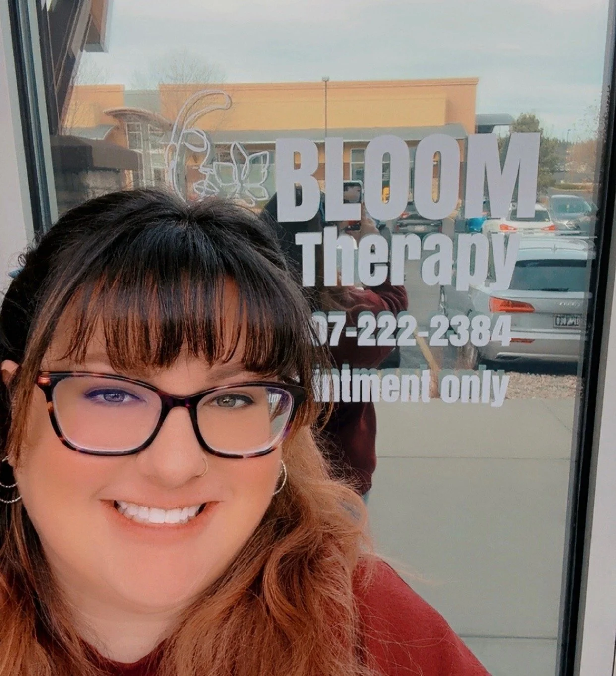 Smiling woman with glasses and earrings standing outside Bloom Therapy storefront with phone number and text, reflected in window.