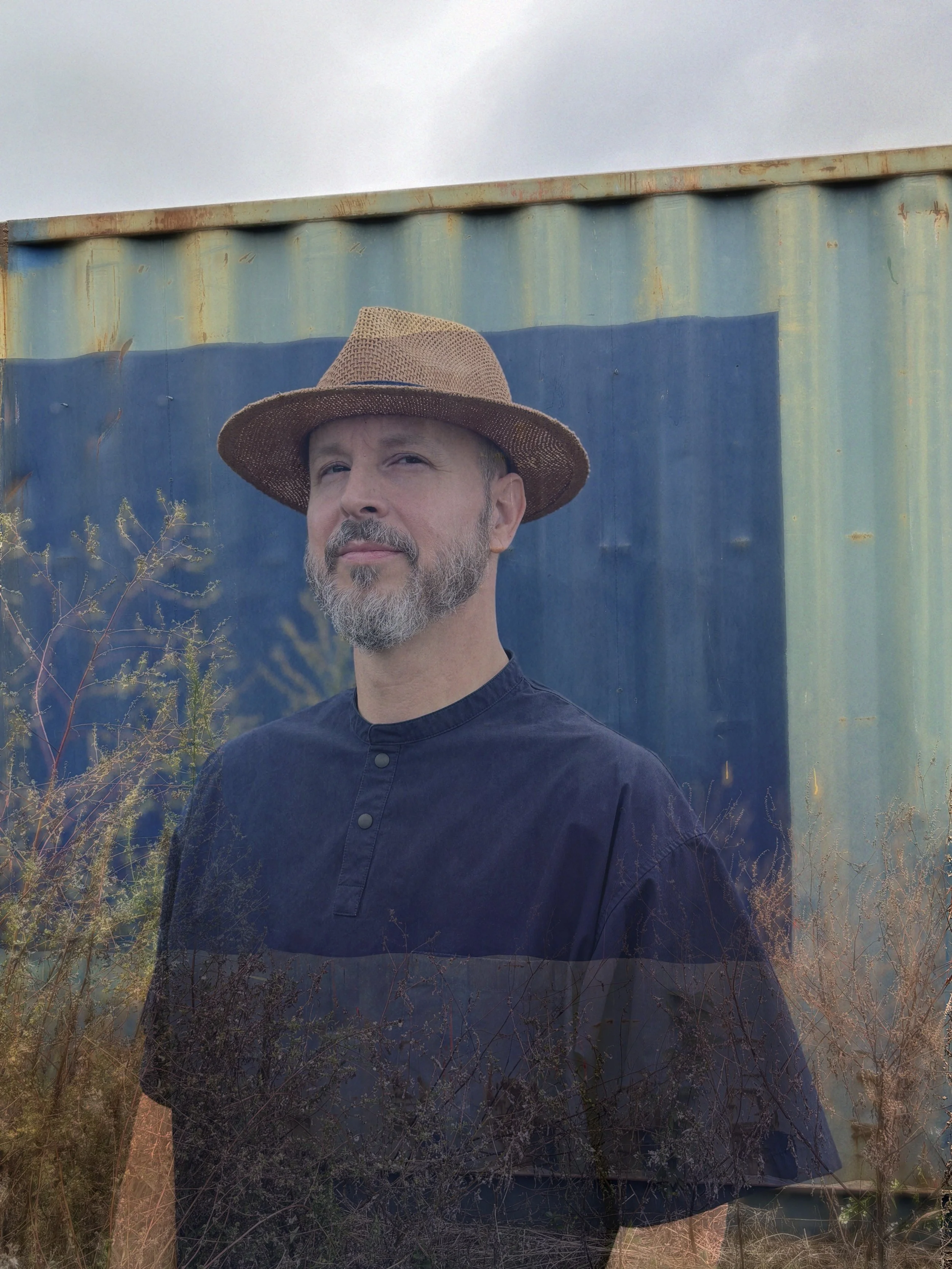 A man with a beard wearing a wide-brimmed hat and a dark shirt, standing outdoors in front of a weathered blue shipping container, with some dry grass and plant life in the foreground.