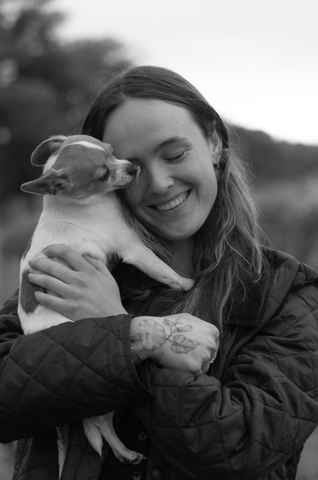 A woman with long hair smiling with eyes closed, holding a small dog close to her face, outdoors.