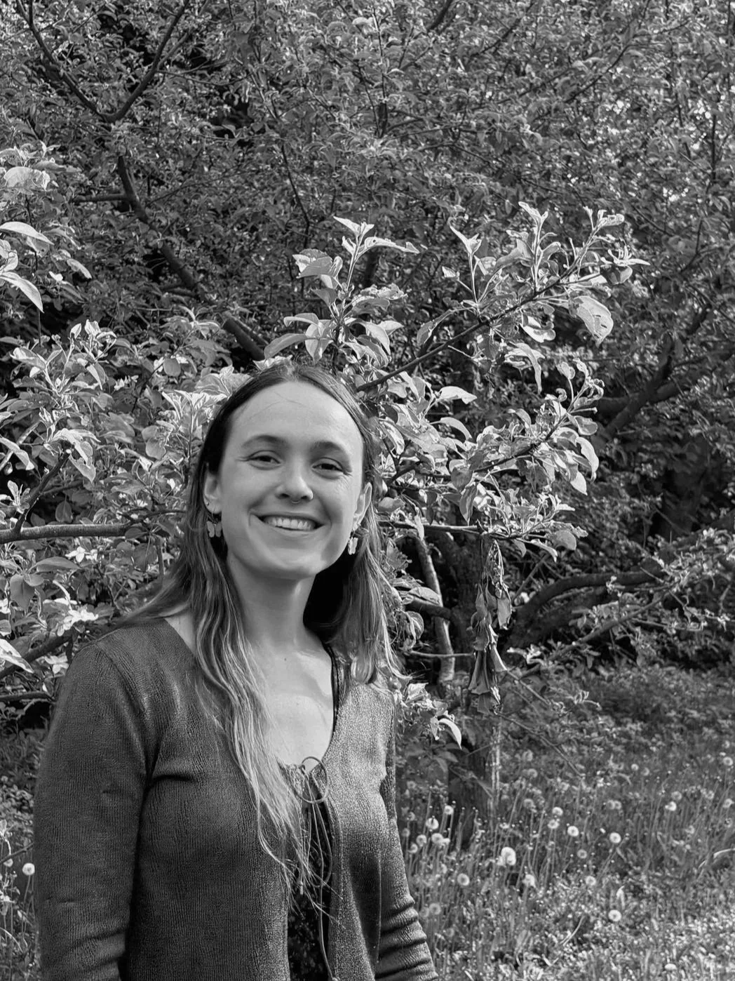 A smiling woman with long hair and earrings stands outdoors in front of a bush and trees.