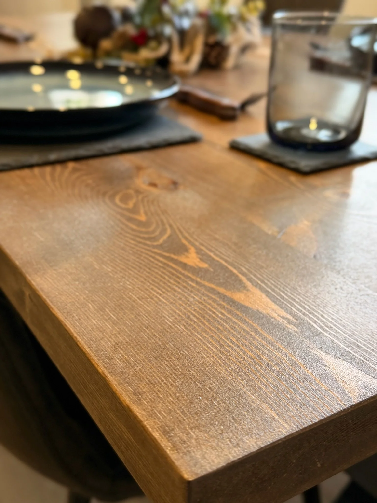 Close-up of a wooden table with a smooth finish, showing the wood grain details and a few tableware items partially visible in the background.