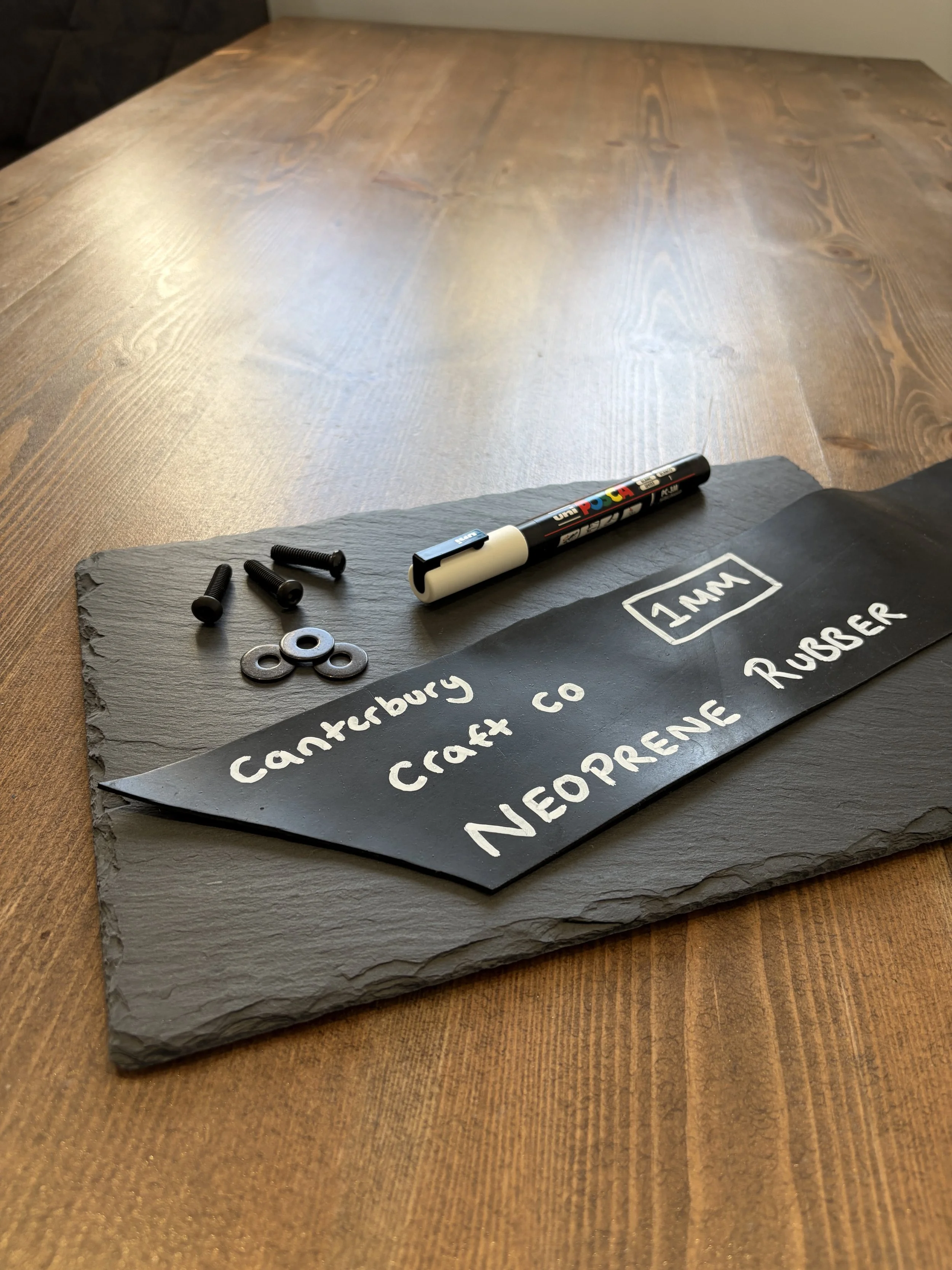 A chalkboard sign on a slate base with handwritten white chalk text mentioning 'Canterbury Craft co' and other text, with black screws and washers placed nearby, on a wooden table.