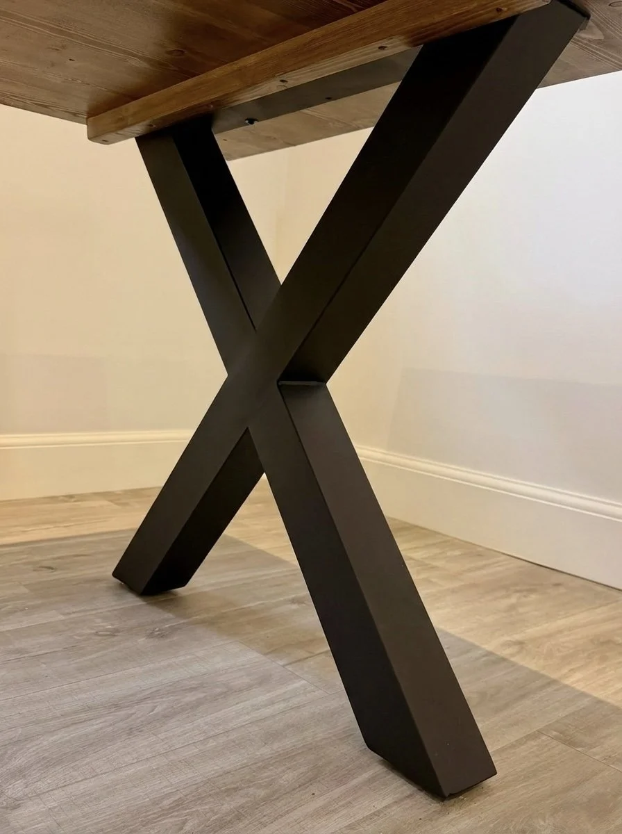 Close-up of a table with a thick wooden top and a unique X-shaped black metal leg base, standing on a wooden floor in front of a plain light-colored wall.