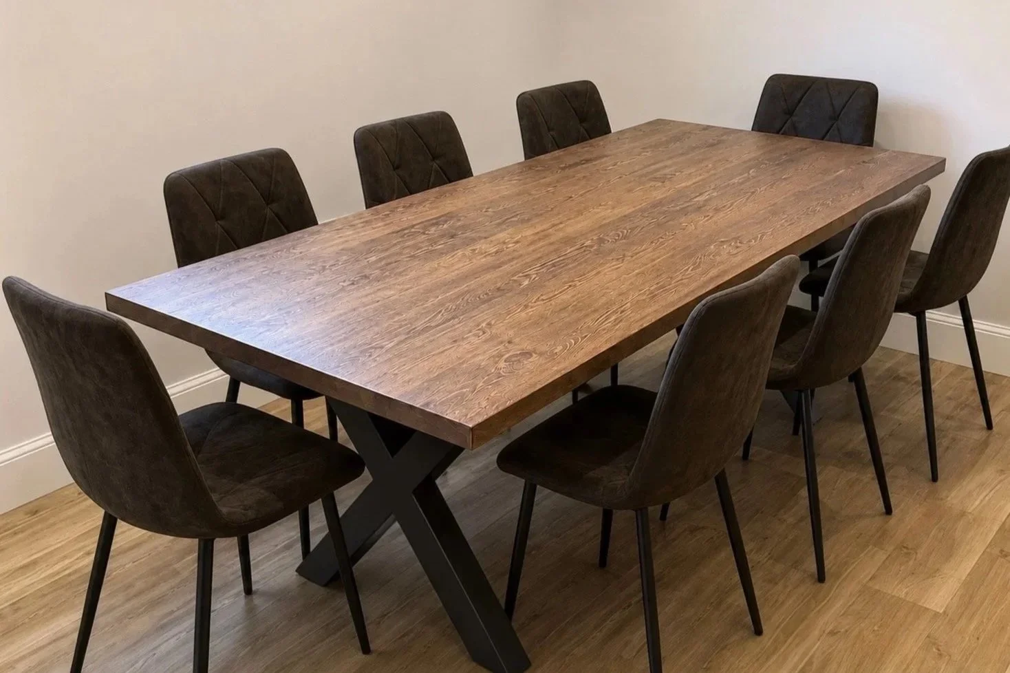 A wooden dining table with a natural finish and black X-shaped legs surrounded by eight dark upholstered chairs in a room with wooden floors and plain beige walls.