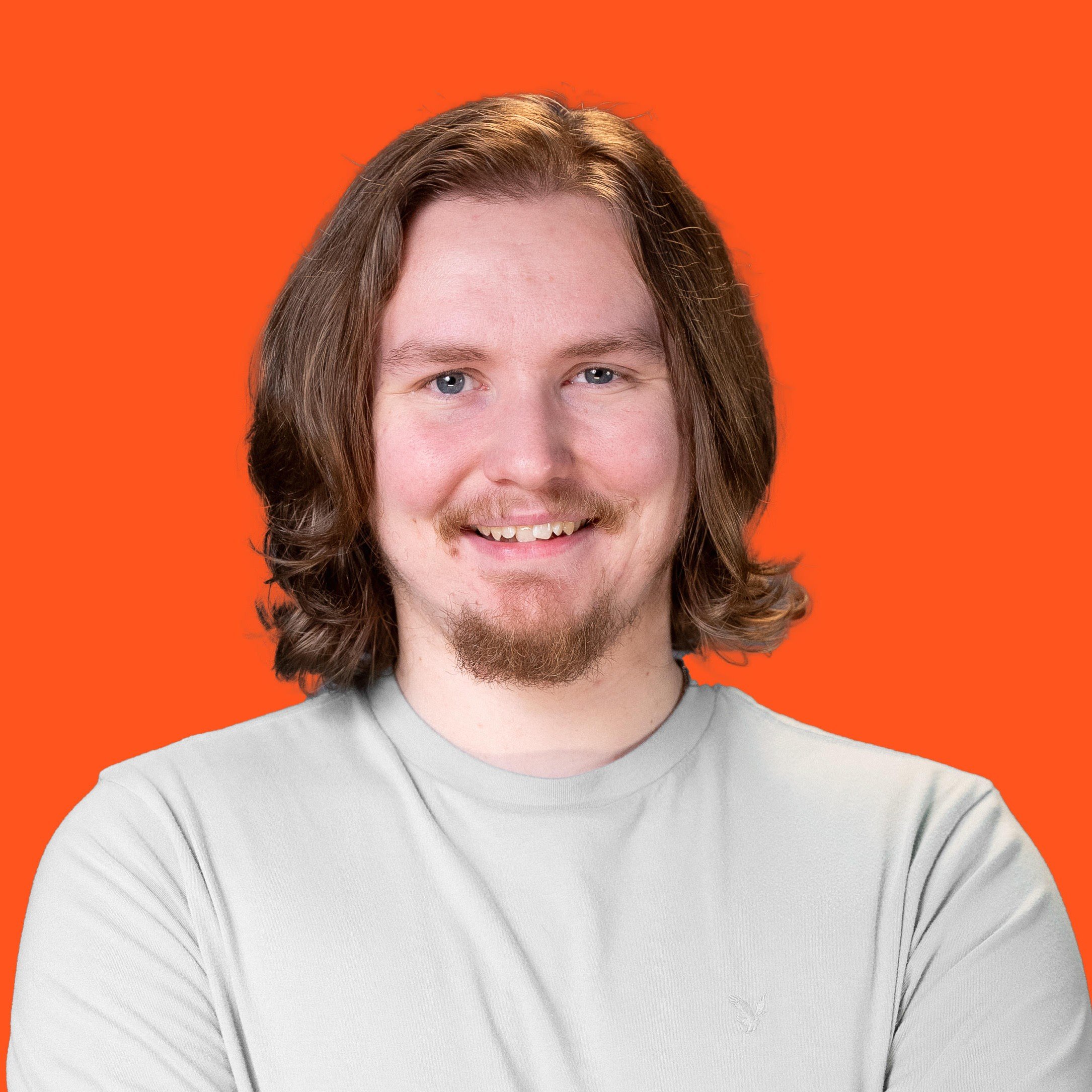Young man with shoulder-length brown hair, light beard, smiling, wearing a light gray shirt, against an orange background.