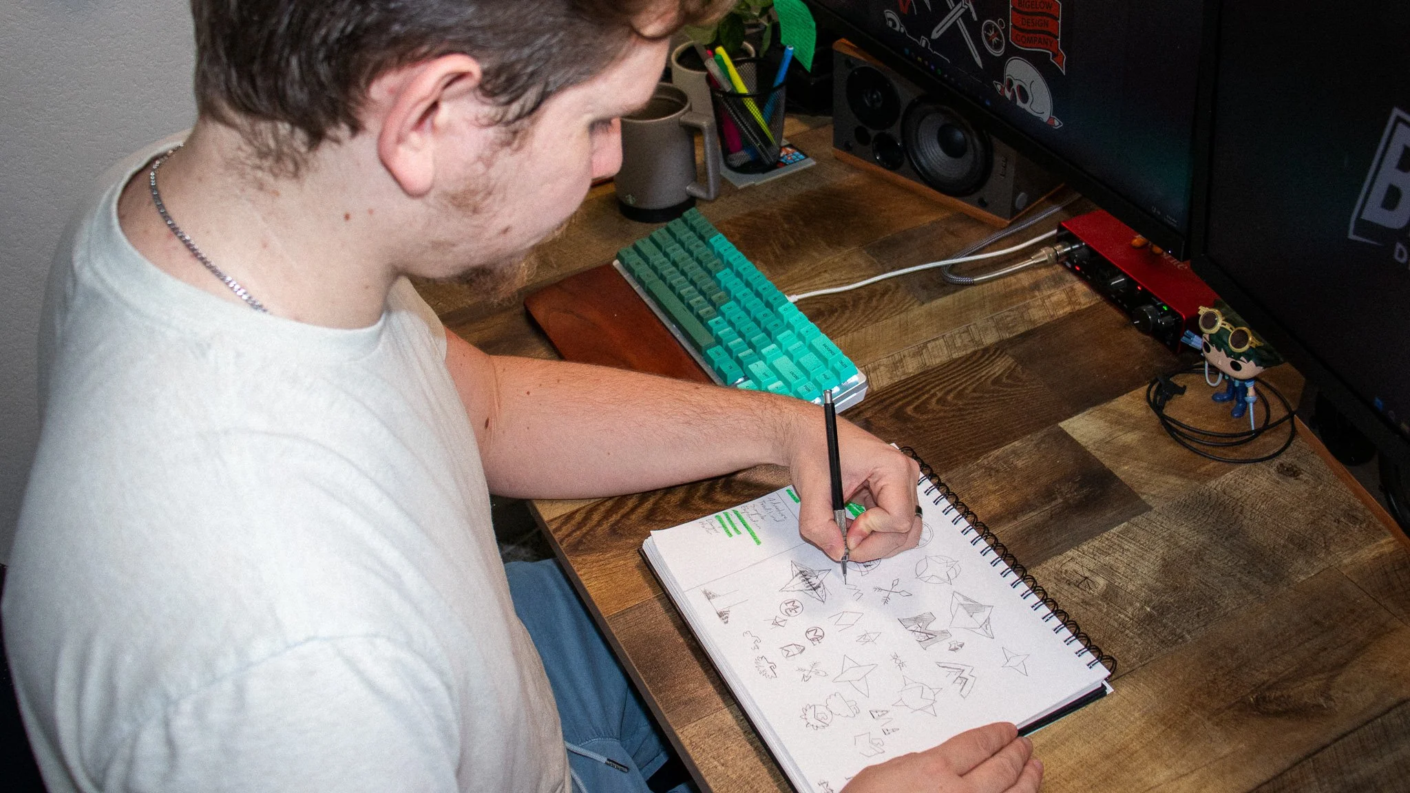A person sitting at a wooden desk writing in a notebook filled with sketches of paper airplanes, with a mechanical keyboard, a monitor, speakers, and various desk accessories in the background.
