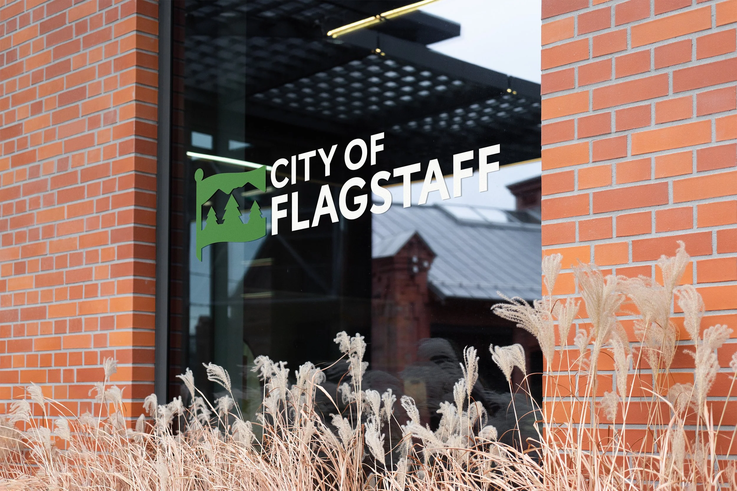 Window with 'City of Flagstaff' logo featuring a green flag with trees, outside brick building, dry ornamental grass in foreground.