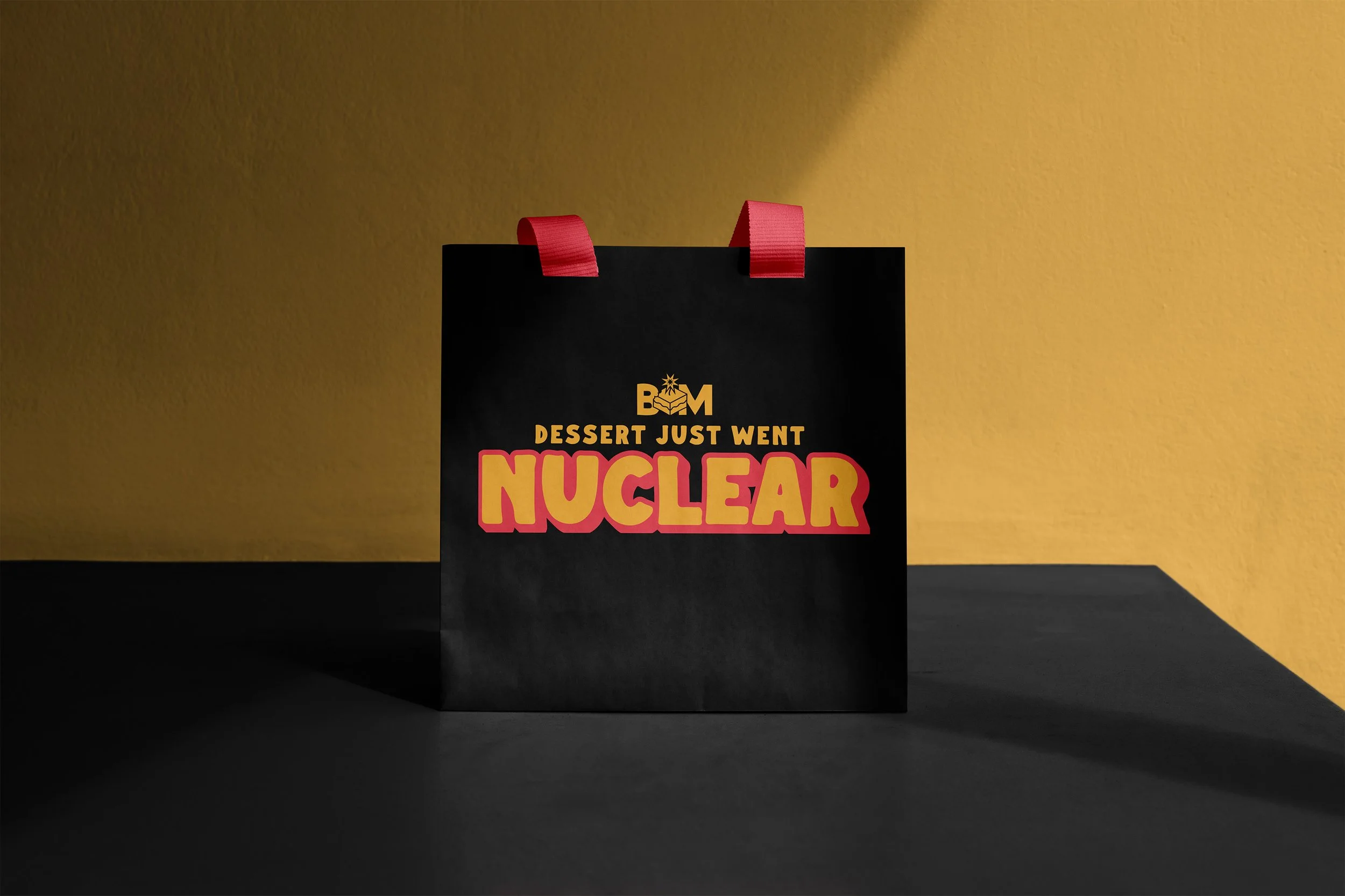 Black shopping bag with red handles, featuring the text 'B*M Dessert Just Went Nuclear' in yellow and pink, against a yellow wall and black surface.