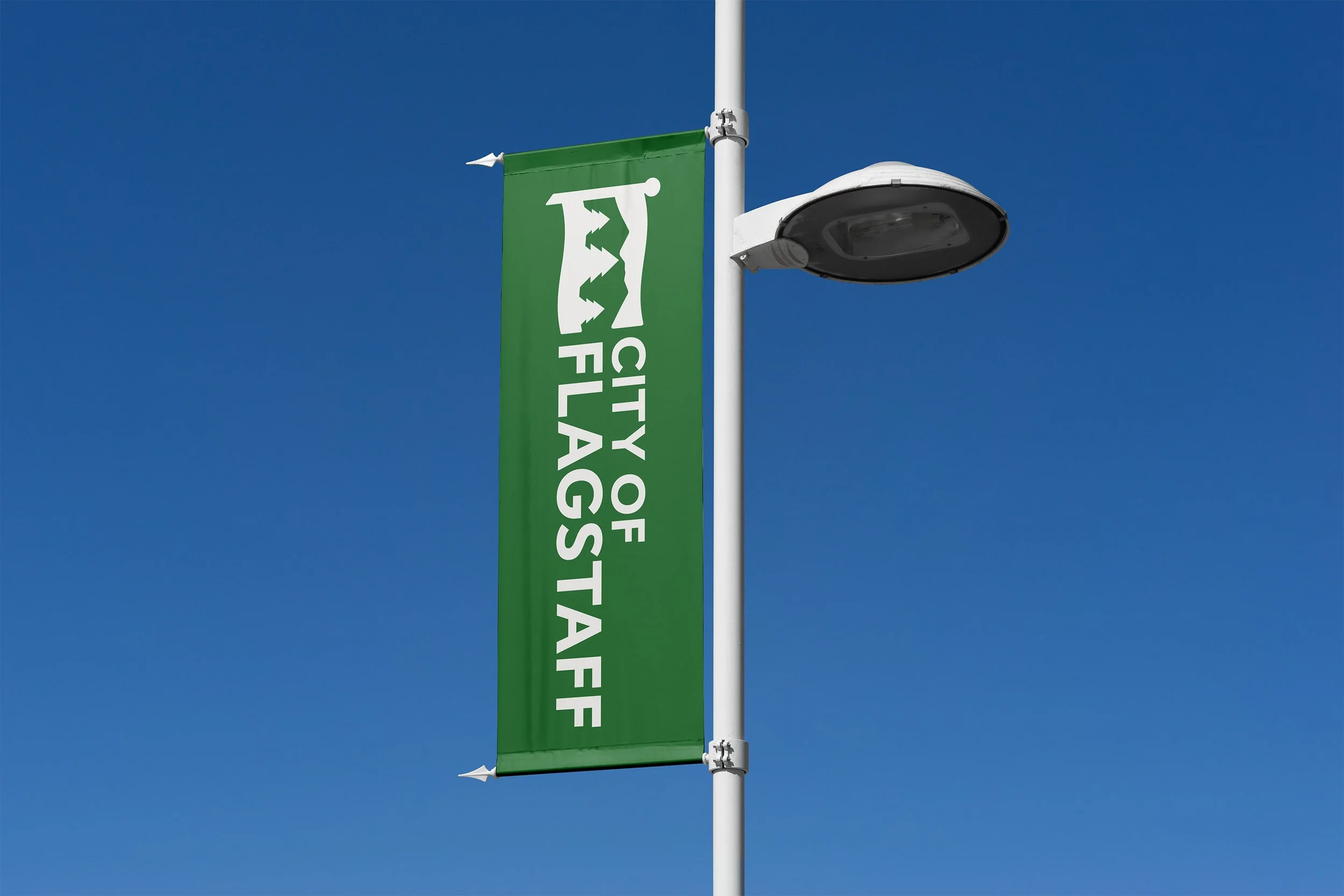 Green flag with white text that says 'City of Flagstaff' and a white emblem of two trees and a mountain, attached to a white pole with a streetlight, against a clear blue sky.