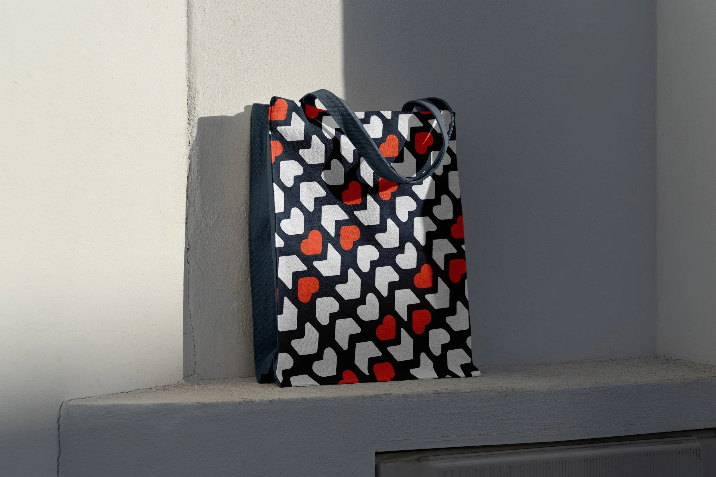 A shopping tote bag with a pattern of white, red, and gray hearts, leaning against a wall in a corner, with sunlight casting a shadow.