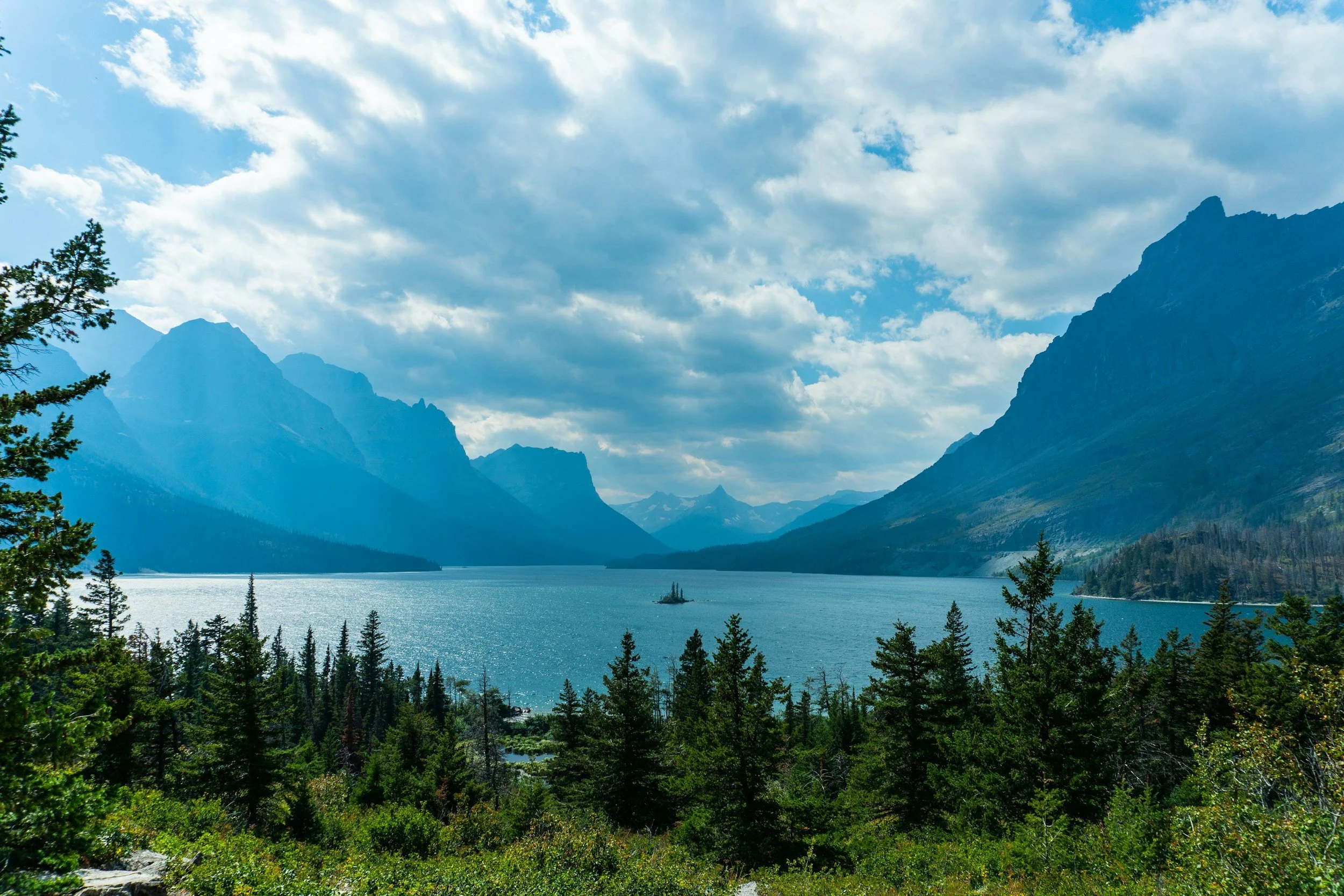 A scenic view of a lake surrounded by mountains and dense forest with a partly cloudy sky.