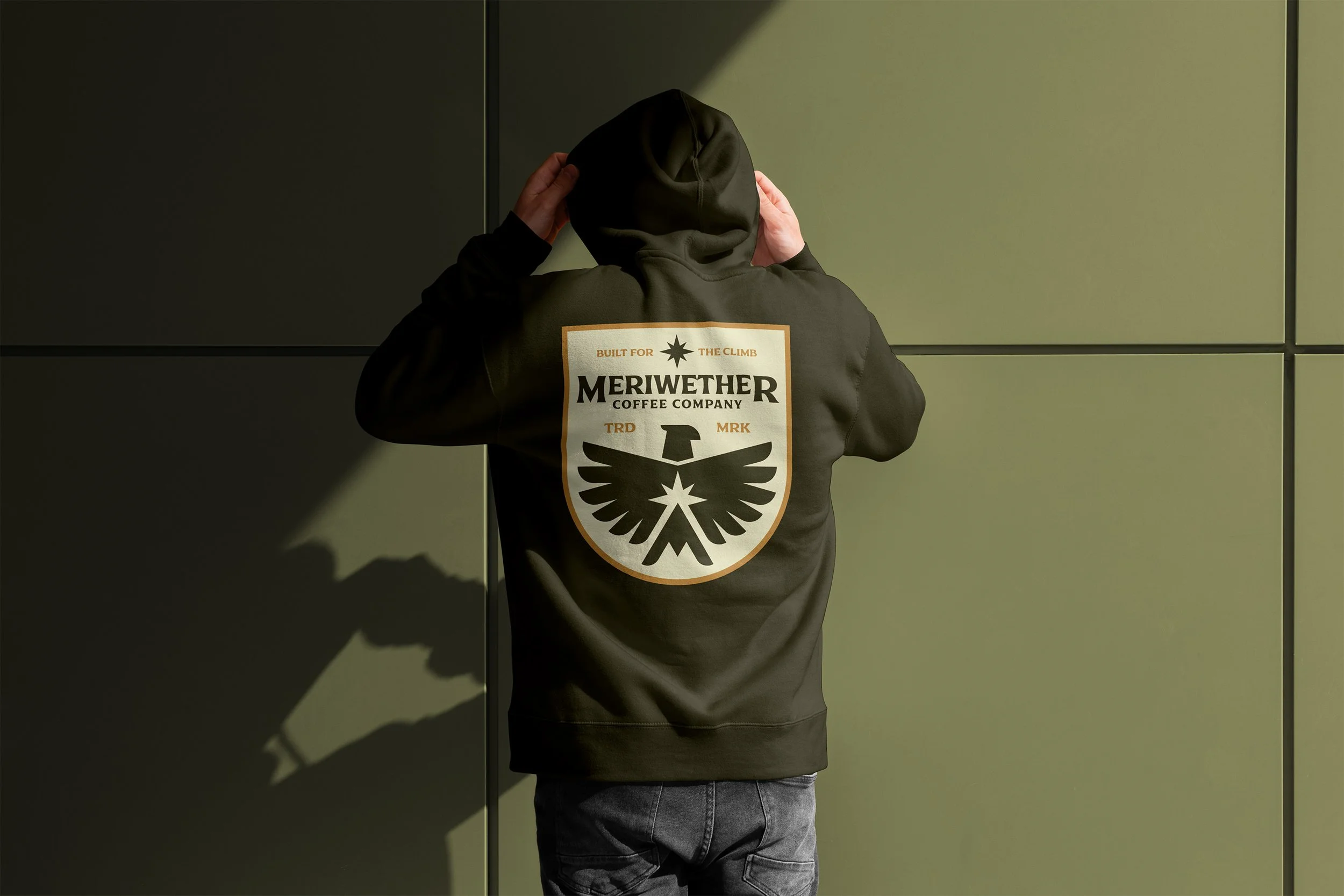Person wearing a hoodie with a large patch on the back reading 'Meriwether Coffee Company' and a logo of an eagle with wings spread, standing against a green wall.