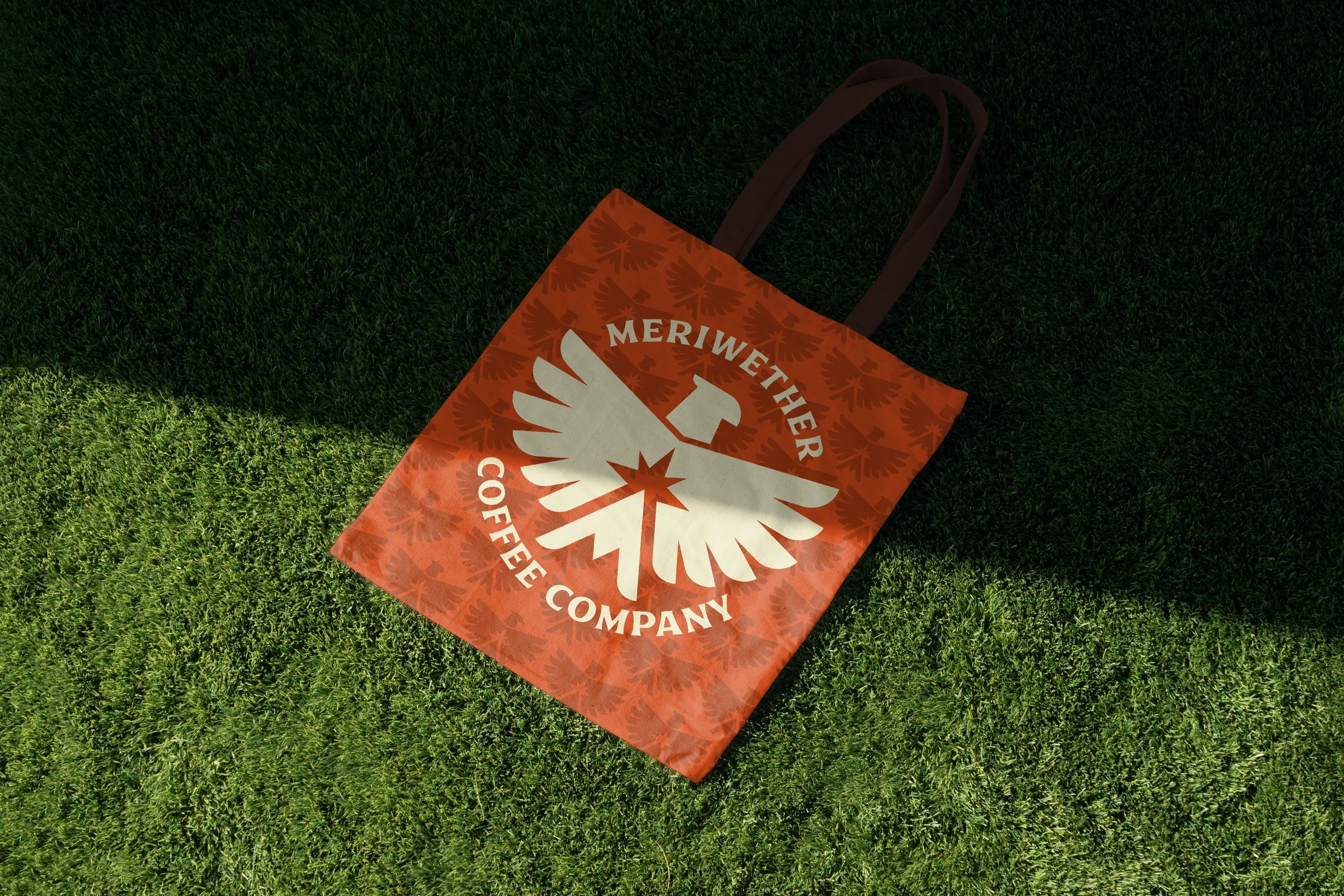 A tote bag with the logo of Meriwether Coffee Company, featuring a white eagle figure with outspread wings and a star on its chest, on an orange background with a pattern of leaves, lying on green grass.