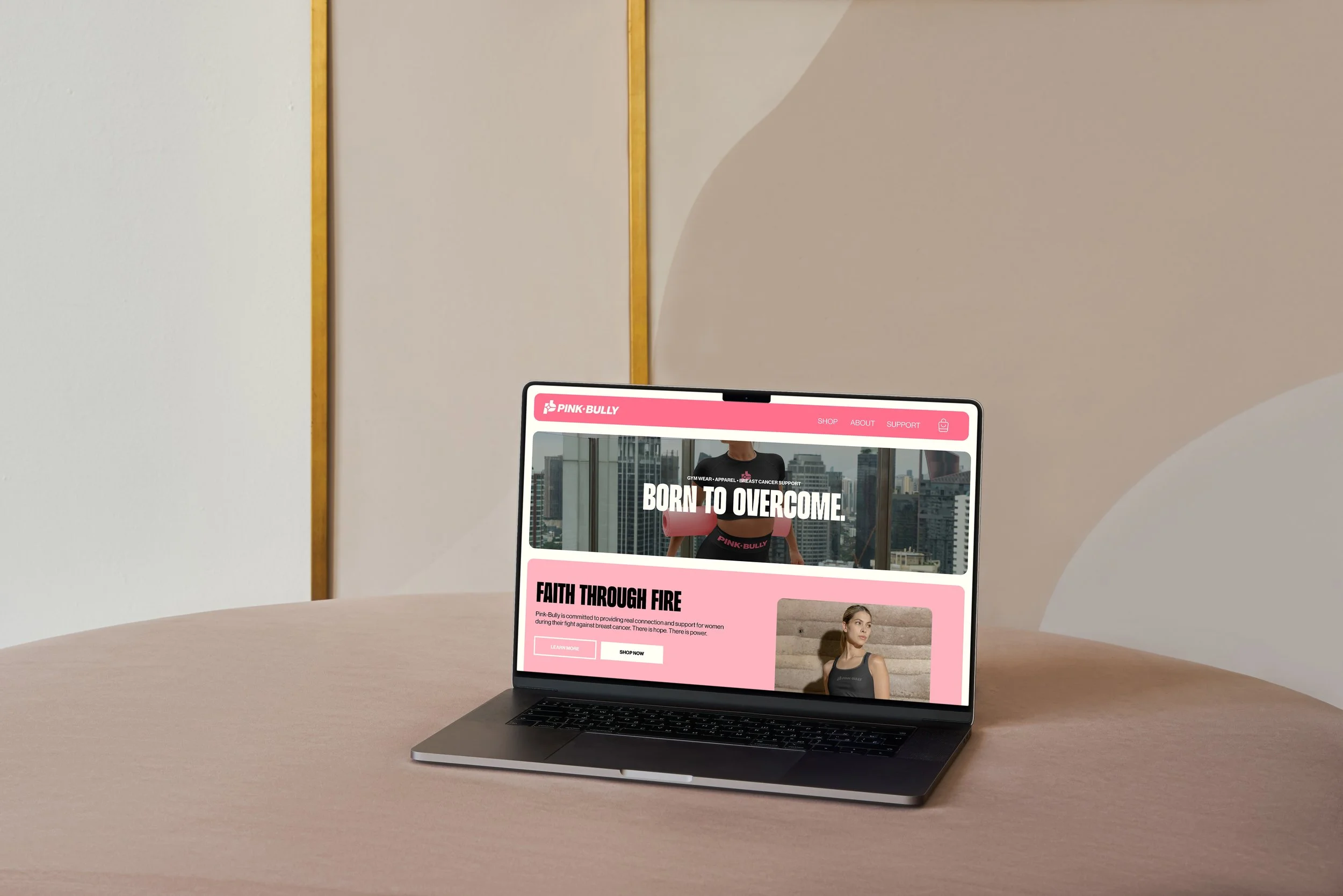 A laptop on a wooden table displaying a pink-themed webpage for Pink Bully, a brand supporting women with breast cancer, featuring an image of a woman in athletic wear and text promoting faith and strength.
