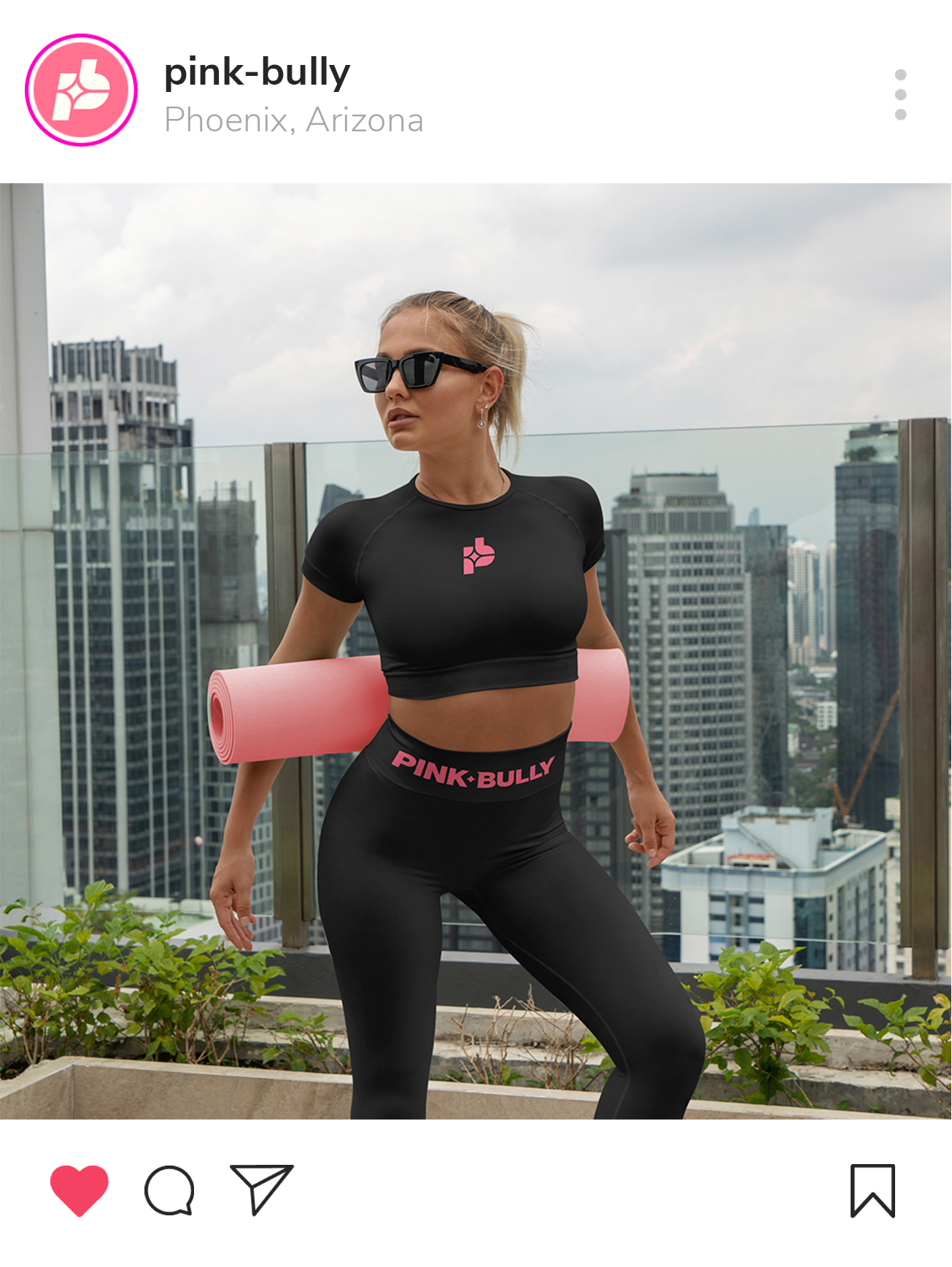 A woman in black PINK BULLY activewear holding a pink yoga mat on her shoulder, standing on a rooftop with a city skyline in the background, wearing sunglasses and jewelry.