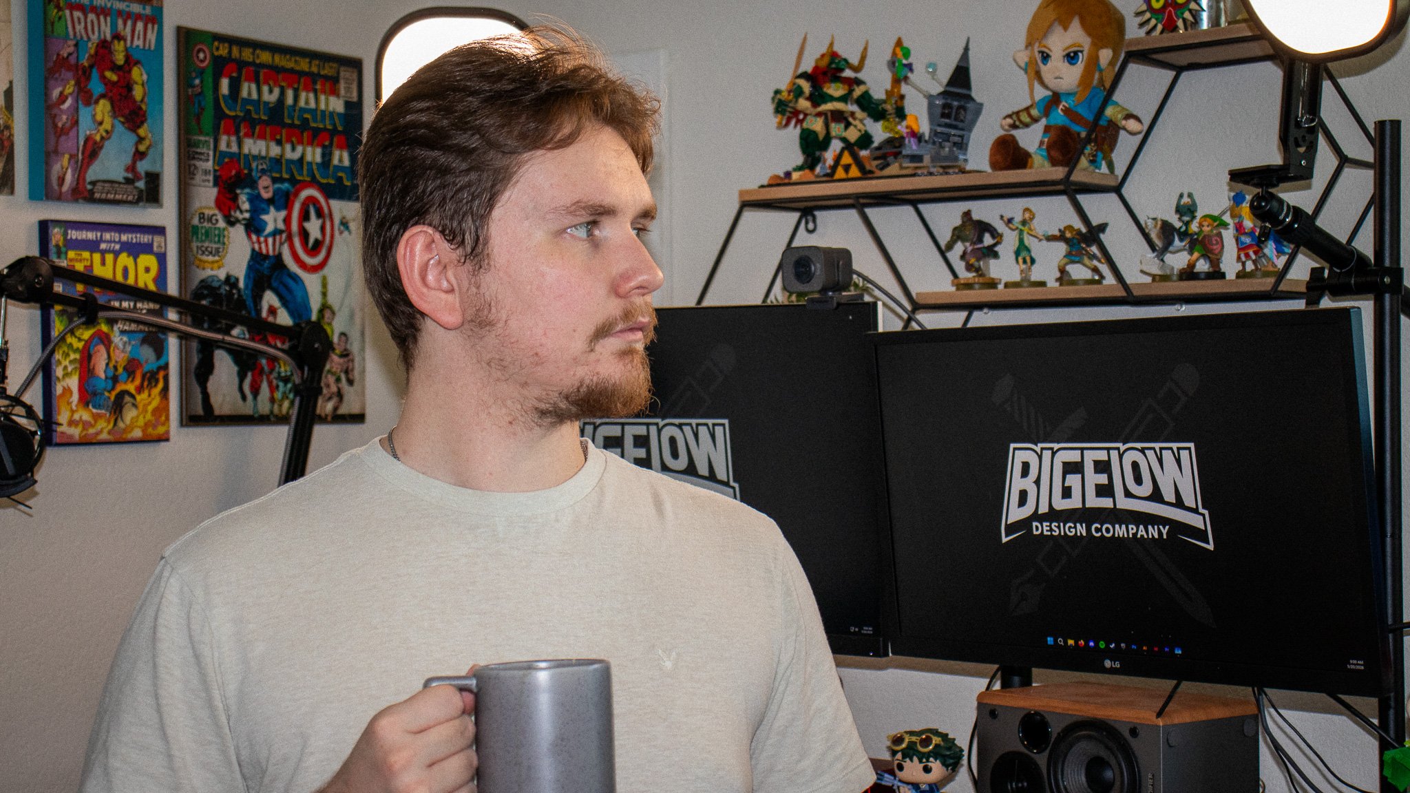 Young man with light brown hair and beard holding a mug, standing in a room decorated with comic book posters, toys, and figurines, with computer monitors and speakers visible.