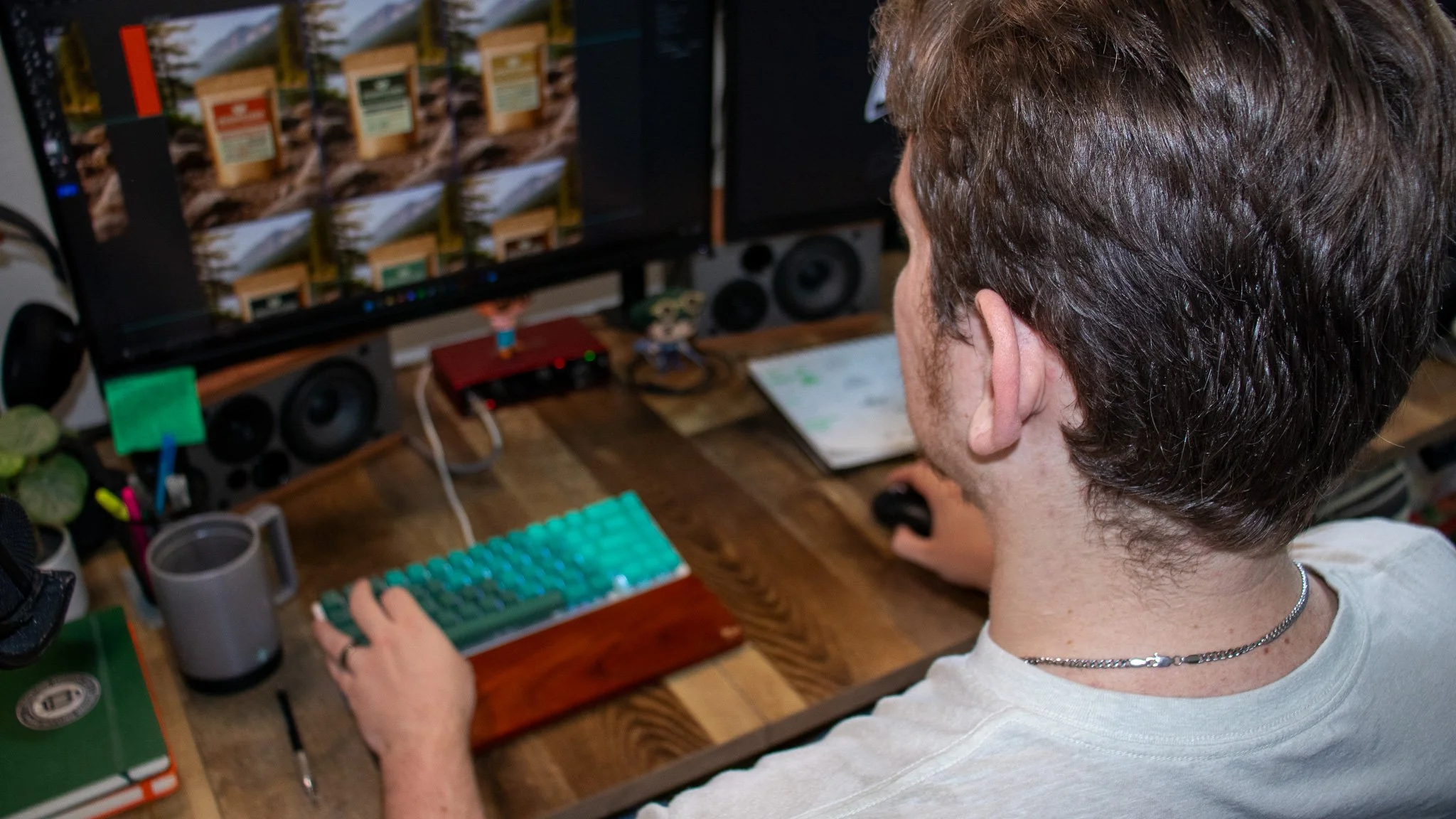 A person with brown hair, wearing a white t-shirt, sitting at a desk working on a computer with multiple screens displaying image editing, keyboard, and other desk items.