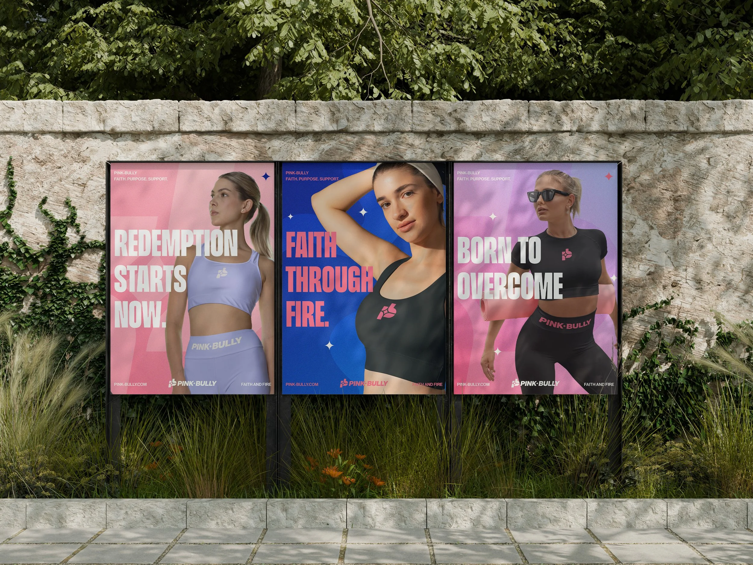 Three large posters on a stone wall featuring women in athletic wear with motivational phrases. The first reads 'Redemption starts now,' the second 'Faith through fire,' and the third 'Born to overcome.' Each woman is styled differently and wearing Pink Bully sportswear.