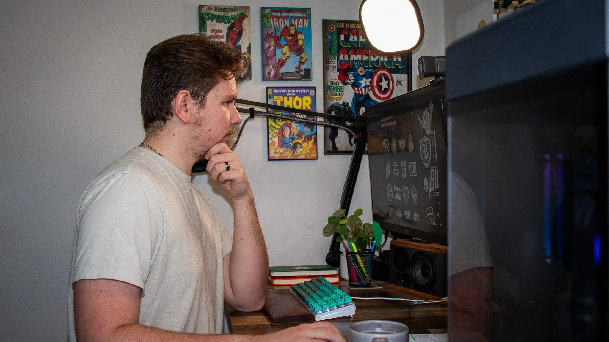 A person with brown hair and a beige T-shirt sitting at a wooden desk, looking at a computer monitor in a home office decorated with comic book posters.