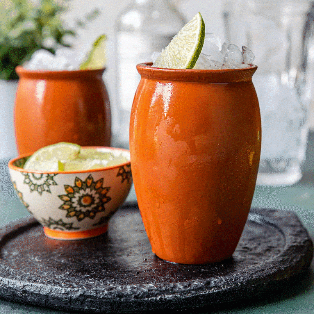 Two orange clay mugs filled with a drink, garnished with lime wedges and ice, placed on a black textured surface.