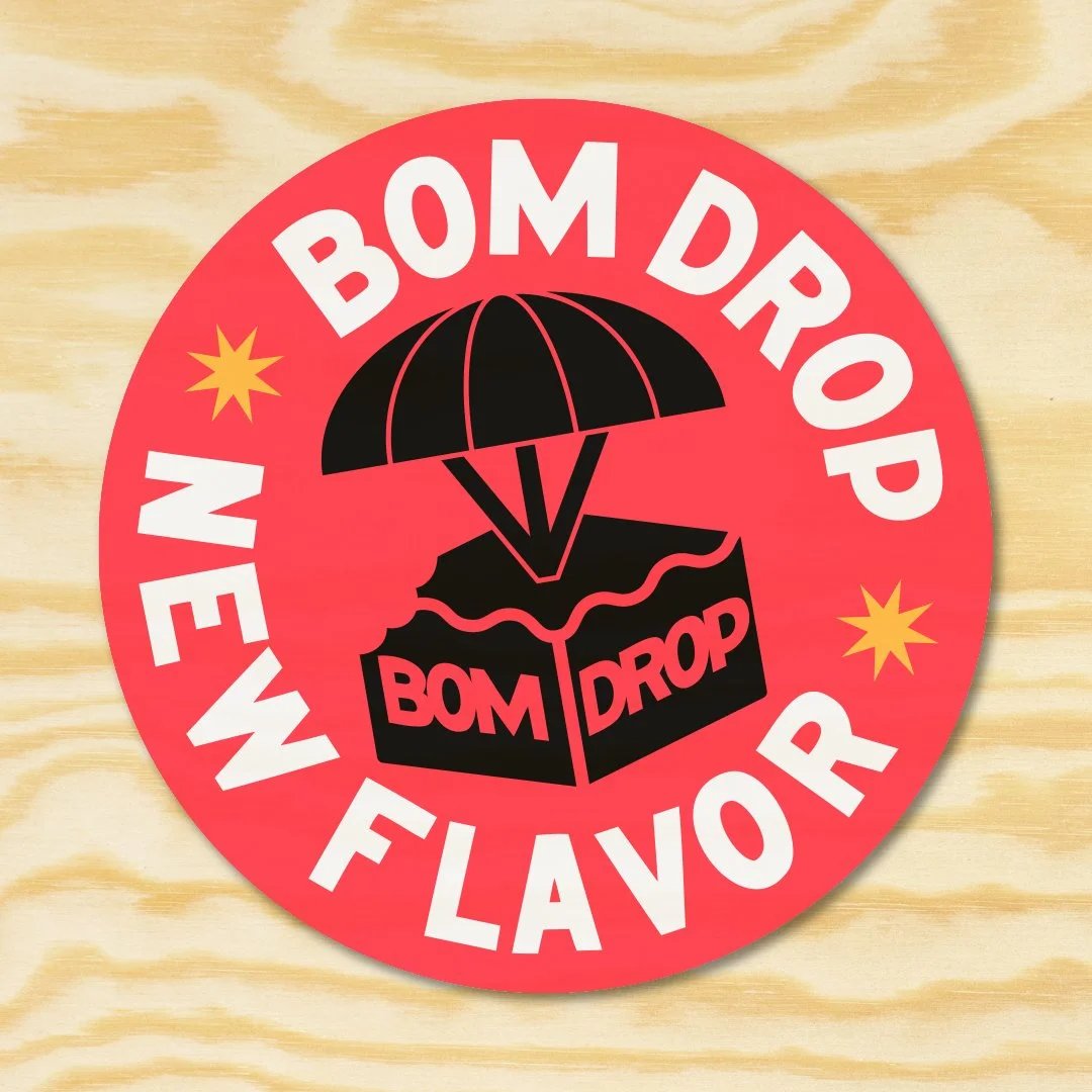 A circular sticker with a red background, featuring a black box with the words 'BOM DROP' and an umbrella above it. The sticker has the text 'NEW BOM DROP' and 'BOM FAVOR' around the circle, with yellow starburst accents.