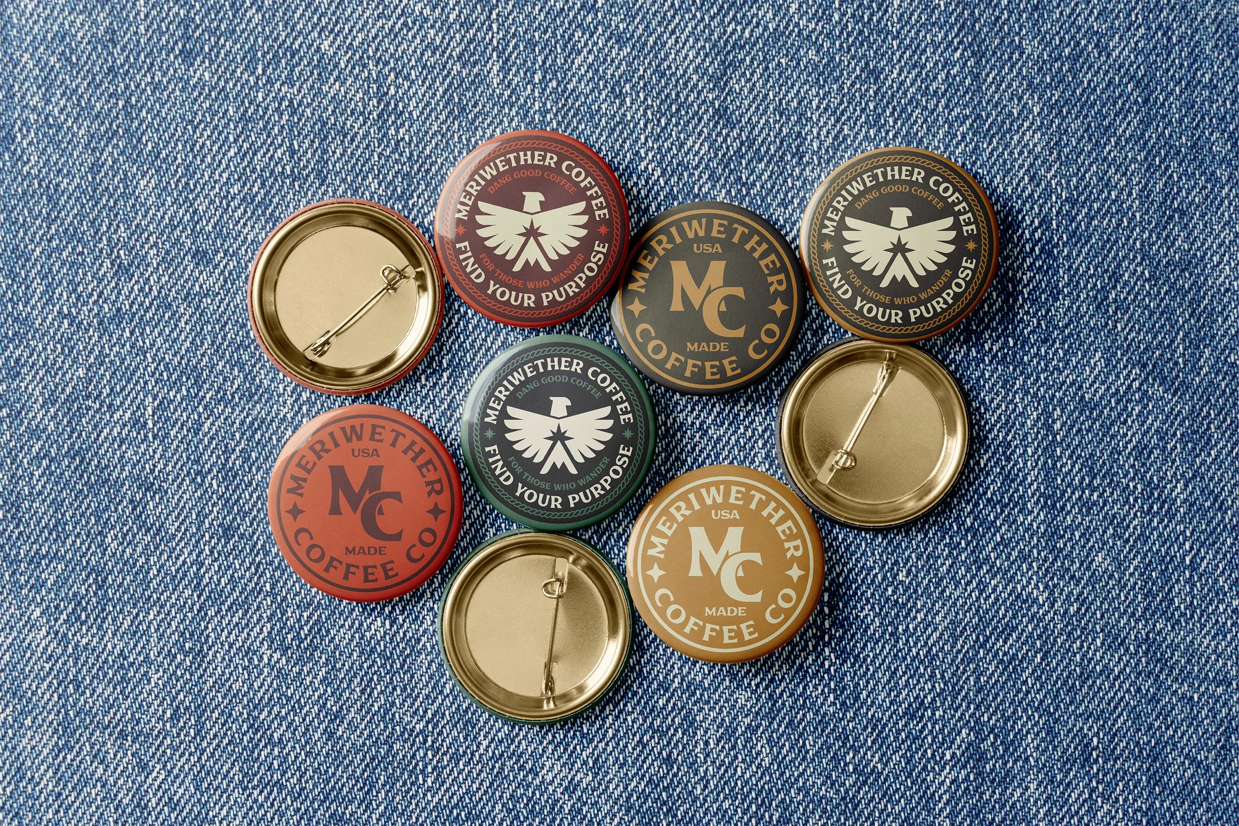 A collection of nine metal pin-back buttons on blue denim fabric, some showing the front design with the 'Meriwether Coffee Co.' logo and others showing the back side with pin fasteners.